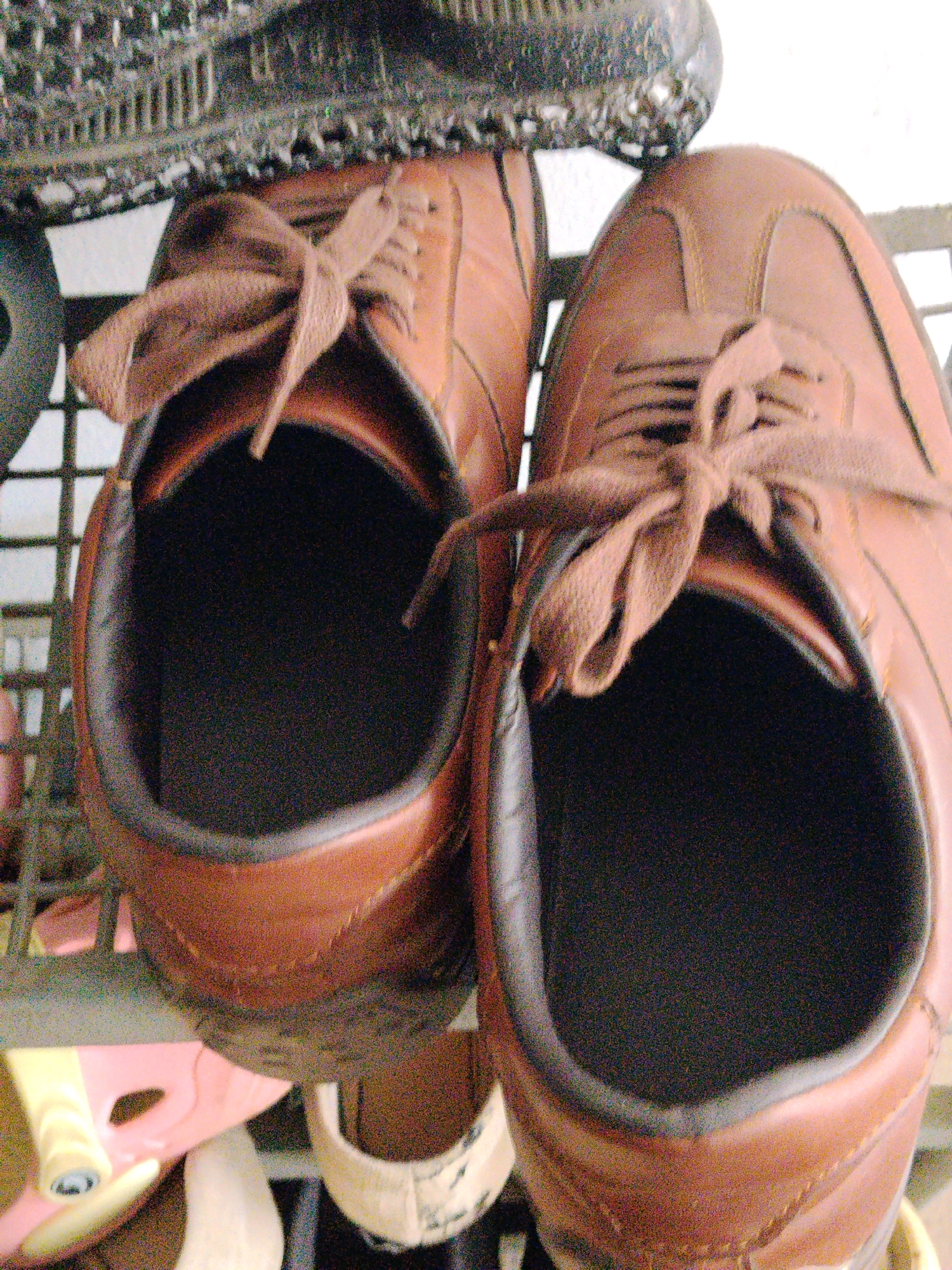 The image displays a close-up, overhead view of two brown lace-up shoes. The shoes are positioned on a rack, with the left shoe slightly angled forward and the right shoe more directly facing the viewer. Both shoes have their laces tied in a bow, and the interior of the shoes, which appears to be black, is visible. In the background, a portion of a black object, possibly another shoe or a shoe rack element, is visible at the top. The overall lighting suggests an indoor or shaded outdoor environment. No specific details indicating the location in Sukabakti, Indonesia are present in the visual information.