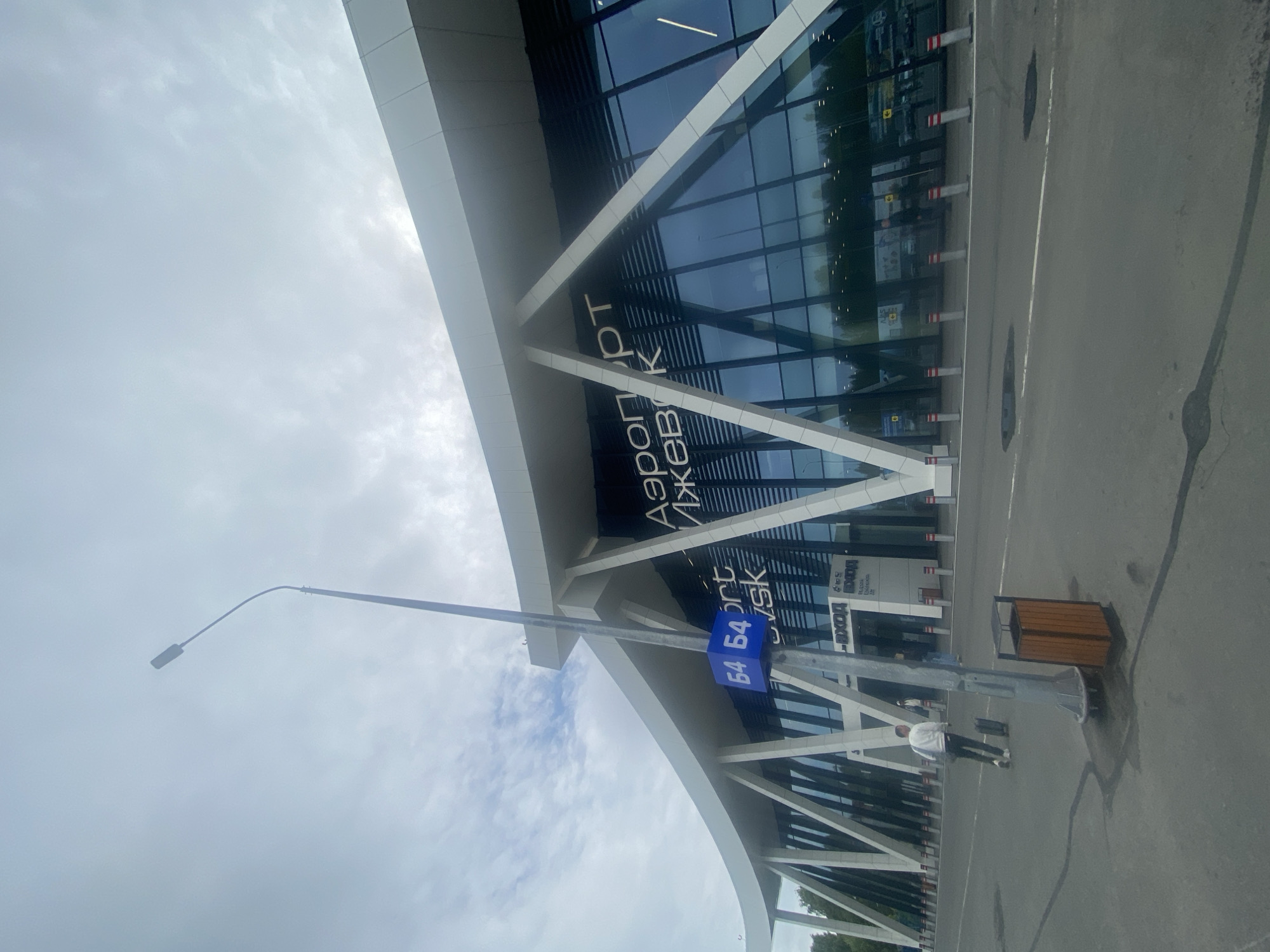 An exterior, low-angle, full shot captures the modern architecture of an airport building under a cloudy sky. The building features a white, curved roof supported by white, angled beams, and large, dark glass windows. The words "Аэропорт Ижевск" (Airport Izhevsk) are prominently displayed in white lettering on the glass facade. To the left of the building, a tall, dark street lamp with a curved arm extends upwards. Attached to the lamp post is a blue sign with white numbers "64" on each side. In the foreground, a man in a white sweatshirt and dark pants is seen from behind, pulling a black rolling suitcase across an asphalt road. A wooden trash receptacle is situated next to the lamp post. The road has white lane markings and several puddles. White and red bollards line the edge of the road in front of the airport entrance, where two other figures are visible near the doors. The environment appears to be an airport arrival or departure area.