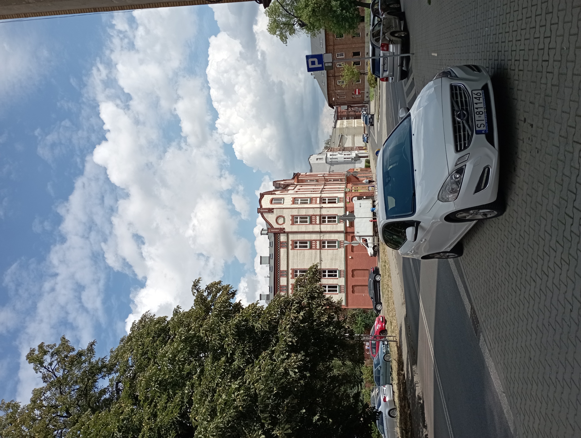 The image depicts a street scene in Siemianowice Śląskie, Poland, on a partly cloudy day. In the foreground, a white Volvo S60 is parked on the right side of the street, facing left. The car's license plate is visible, reading "SI 81146". Adjacent to the car is a cobblestone sidewalk. Across the street, several other cars are parked along the curb, including a silver sedan and a red hatchback. Behind the parked cars, buildings with red brick and cream-colored facades line the street. A large, leafy green tree is prominent on the left side of the image, casting shadows on the sidewalk and street. The sky is a bright blue with scattered white clouds. A blue parking sign with a white "P" is visible further down the street. The overall scene suggests a typical urban environment on a pleasant day.