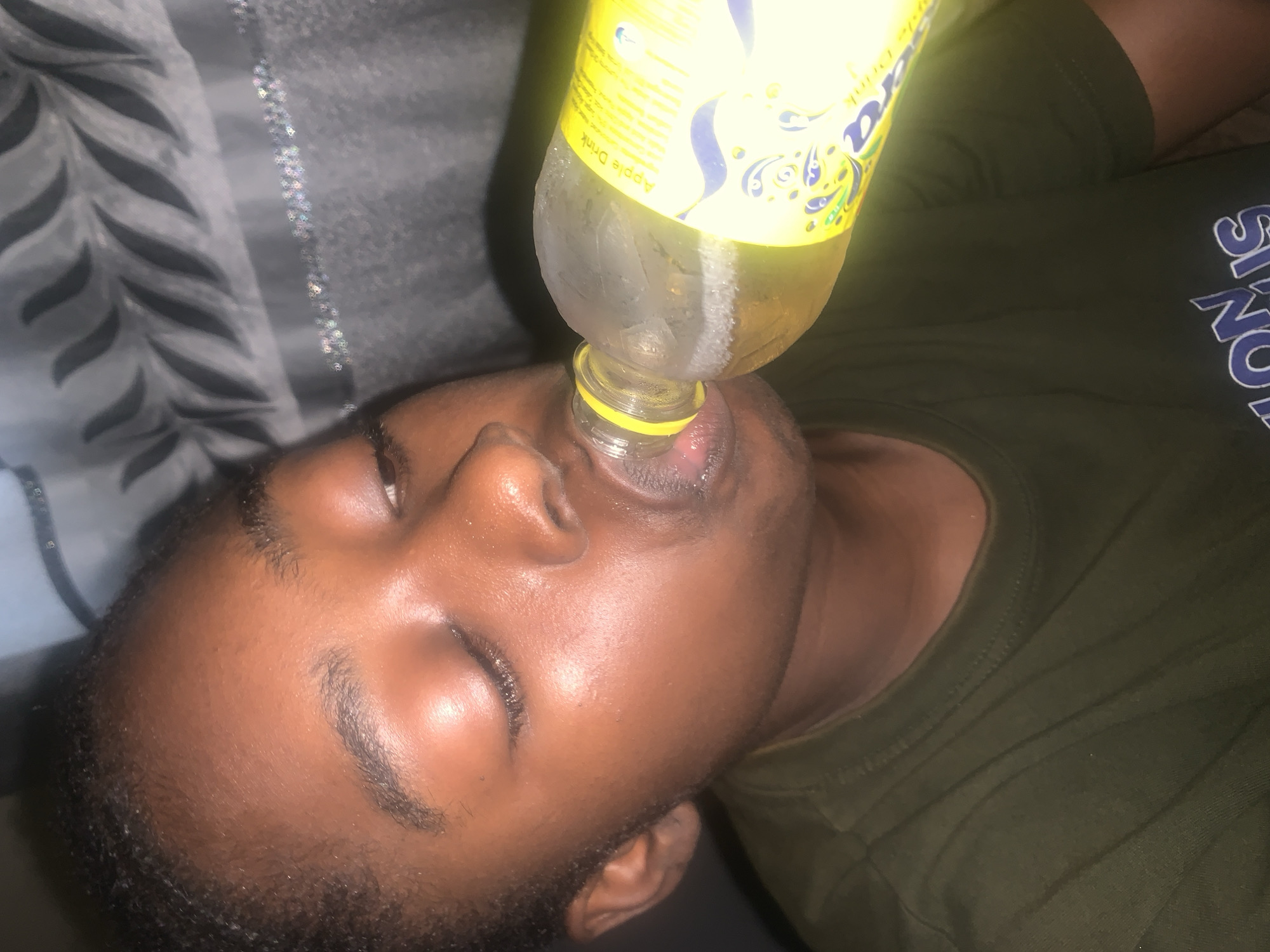 A person lies on a bed and drinks from a bottle. The person is of African descent and has dark skin and short, dark hair. The person is wearing a green shirt. The bottle is yellow and contains a liquid that appears to be apple juice. The bed is covered with a grey and white patterned sheet. The location is in Rafin Par, Nigeria.