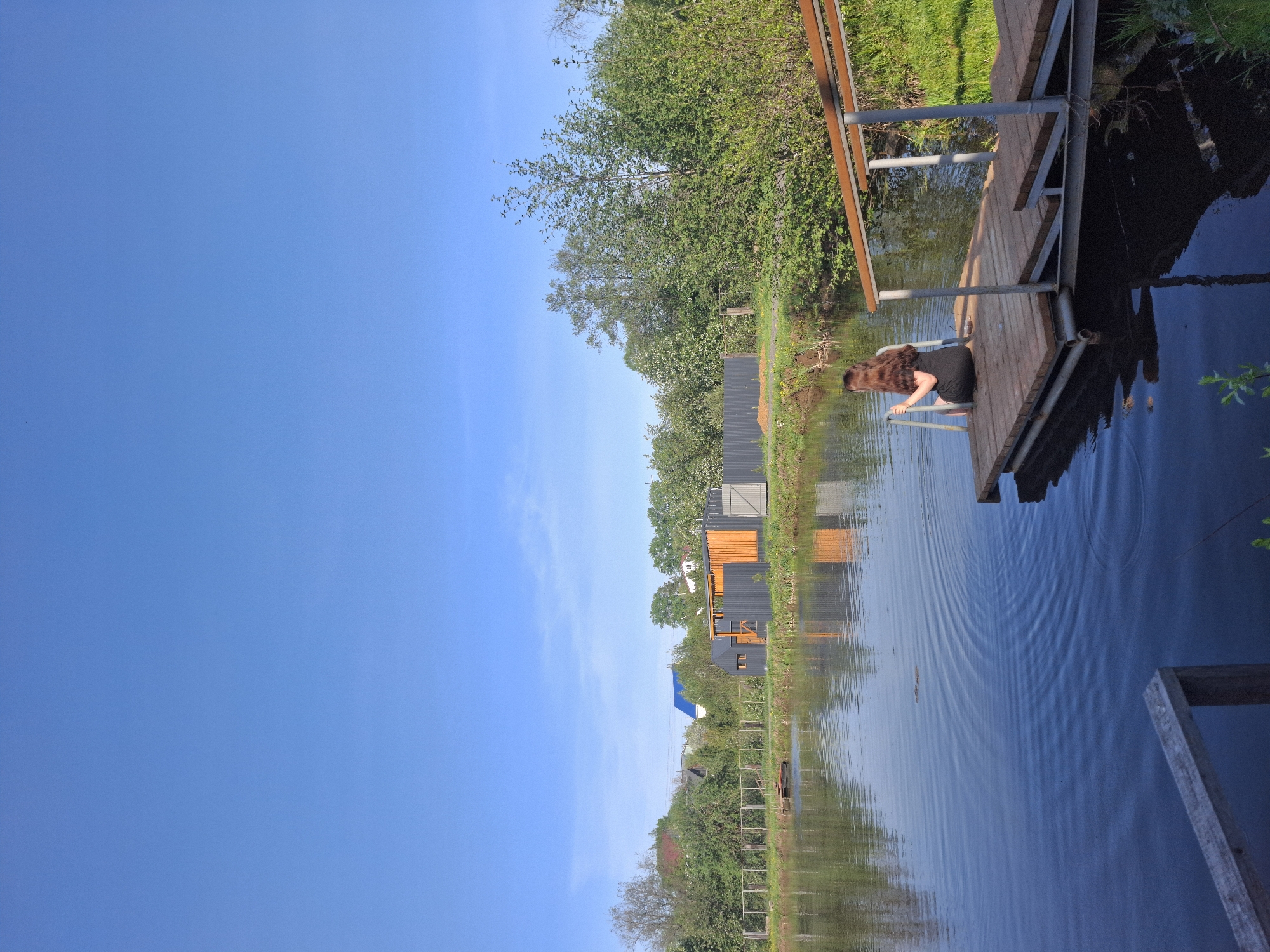 The photograph depicts a sunny, clear day in Malyi Bukor, Russia. A body of water, likely a river or lake, stretches across the majority of the scene, reflecting the blue sky and surrounding structures. The reflections include buildings on the shoreline, some with wooden facades and others with more modern, dark siding and orange accents. Vegetation lines the banks of the water, with trees and grasses visible. A wooden dock extends out over the water, and a person with long, brown hair is positioned on the edge of the dock, near a ladder that leads into the water. The person is wearing a dark dress or shirt and appears to be getting ready to enter the water. The water's surface shows ripples, indicating movement or disturbance.