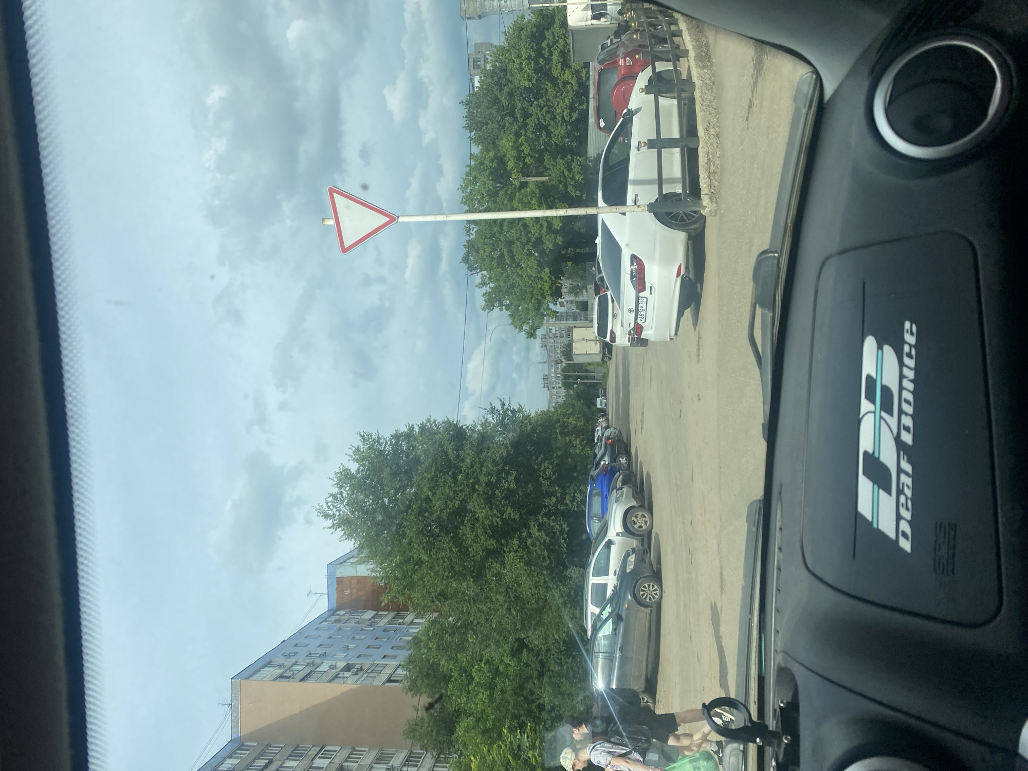 The image, taken from inside a car in Samara, Russia, shows a street scene under a cloudy sky. A yield sign is prominently displayed on a white pole in the left portion of the frame. Several cars are parked along the side of the road, including a white sedan with a license plate visible, a blue SUV, and other vehicles further down the street. Lush green trees line the road, with apartment buildings visible in the background. The dashboard of the car is partially visible on the right, featuring a circular air vent and a sticker with the text "DEAF DONCE". Two people are partially visible in the foreground, appearing to be walking near the parked cars. The overall impression is a daytime scene, likely in a residential or commercial area, with overcast weather.