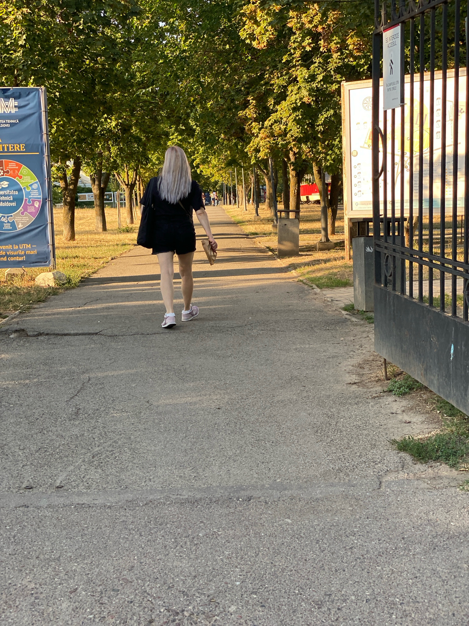 A full shot captures a young woman with blonde hair walking away from the camera on a paved path at the entrance of a university campus. She is wearing a black t-shirt, black shorts, and pink sneakers. She is carrying a brown paper bag in her right hand. The path is lined with trees on both sides. On the left side of the path, there is a blue banner with text and graphics. On the right side, a black metal gate is partially open, revealing a sign with a map of the campus and a smaller sign indicating that access is restricted. The sun is casting long shadows across the path, suggesting it is late afternoon or early evening. The environment appears to be a park-like setting within an urban area, consistent with the surroundings of the Technical University of Moldova in Chișinău, Moldova. The overall scene conveys a sense of movement and transition as the woman enters or exits the university grounds.