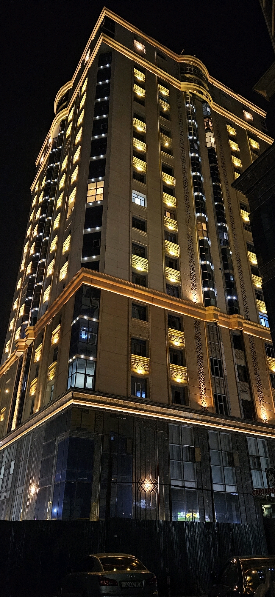 The image depicts a brightly lit, modern high-rise building at night in Dushanbe, Tajikistan. The building features a light beige facade with numerous illuminated windows, some emitting a warm yellow glow and others a cooler white light. Decorative elements, such as ornate carvings and illuminated bands of light, accentuate the architectural features of the building, particularly around the windows and along the horizontal levels. The lower section of the building is characterized by a glass facade, reflecting the surrounding lights. In the foreground, a dark metal fence obscures the view of the ground level, with two cars partially visible behind it. One car, a silver sedan, is positioned in the lower left of the frame, displaying a license plate with Cyrillic characters and the inscription "02002 JD3". The overall scene suggests an urban setting at night, with the building being a prominent and illuminated structure.