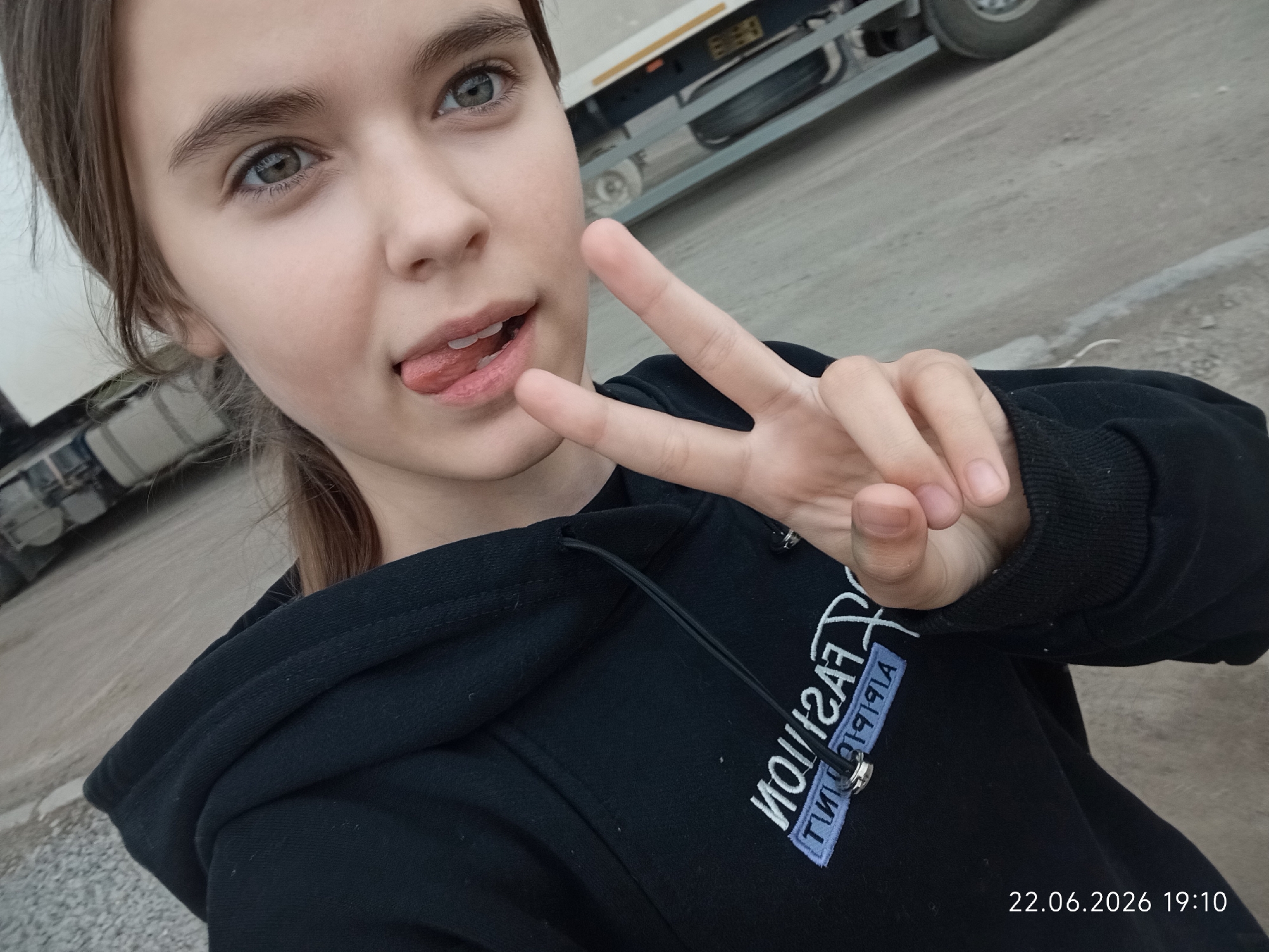 A young female with light brown hair is taking a selfie outdoors, with a truck in the background. She is sticking her tongue out slightly and holding up a peace sign with her right hand. She is wearing a black hoodie with white lettering on it that reads "Fashion" in a stylized font. The date and time stamp on the image reads "22.06.2026 19:10". The background appears to be a dirt or gravel lot with a paved surface and some construction equipment visible. The weather appears to be overcast. The location is specified as Sarytogay, Kazakhstan.