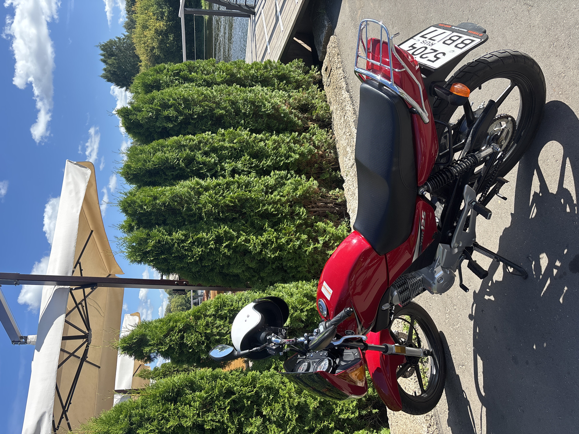 A red Yamaha motorcycle with a white helmet resting on its handlebars is parked outdoors on an asphalt surface. The motorcycle is positioned in front of a row of tall, green evergreen trees. In the background, a body of water with a wooden deck and a railing is visible. The sky is blue with scattered white clouds. The scene appears to be in Zvenigorod, Russia, possibly near a cafe or restaurant with outdoor seating indicated by large, beige umbrellas. The license plate on the motorcycle reads "5204 BB 77".