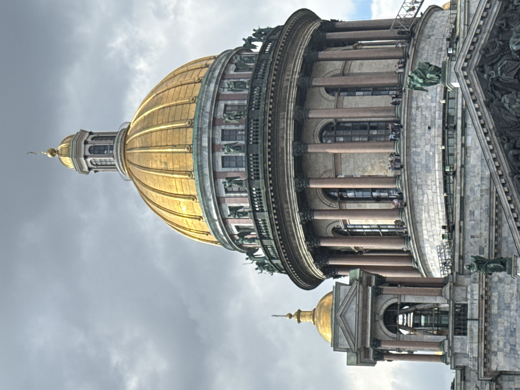 This image captures a close-up view of the golden dome and upper structure of St. Isaac's Cathedral, a prominent landmark in Saint Petersburg, Russia. The sky above is overcast with dramatic, dark clouds. The cathedral's architecture is intricate, featuring a large, gleaming golden dome topped with a smaller cupola and cross. Around the base of the main dome, there is a circular colonnade with statues of angels adorning the balustrade. Below this, a facade with large arched windows and classical columns is visible. To the left, a smaller structure with a golden dome and a bell tower-like element is also visible. Several people are present on a balcony and walkway encircling the main dome, appearing as small figures against the grand scale of the building. Another statue of an angel is positioned on a lower level of the cathedral. The stonework of the building is detailed, with a mix of smooth and textured surfaces. The overall impression is one of architectural grandeur and a slightly dramatic, cloudy atmosphere.