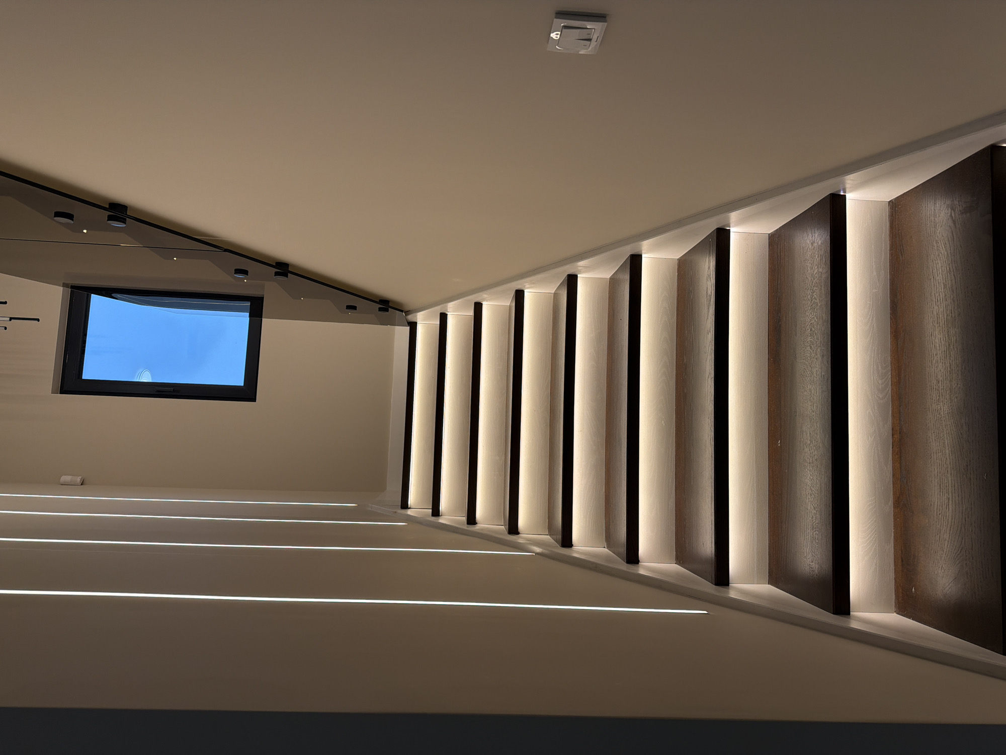 The image shows an indoor staircase illuminated with built-in LED strip lights along the wall and underneath the steps. A rectangular window is visible in the upper left corner. The staircase has dark wood panels alternating with illuminated white sections. The walls and ceiling are light beige. There is a square-shaped object on the ceiling, possibly a smoke detector or sensor. The location is unknown. There are no people in the image.