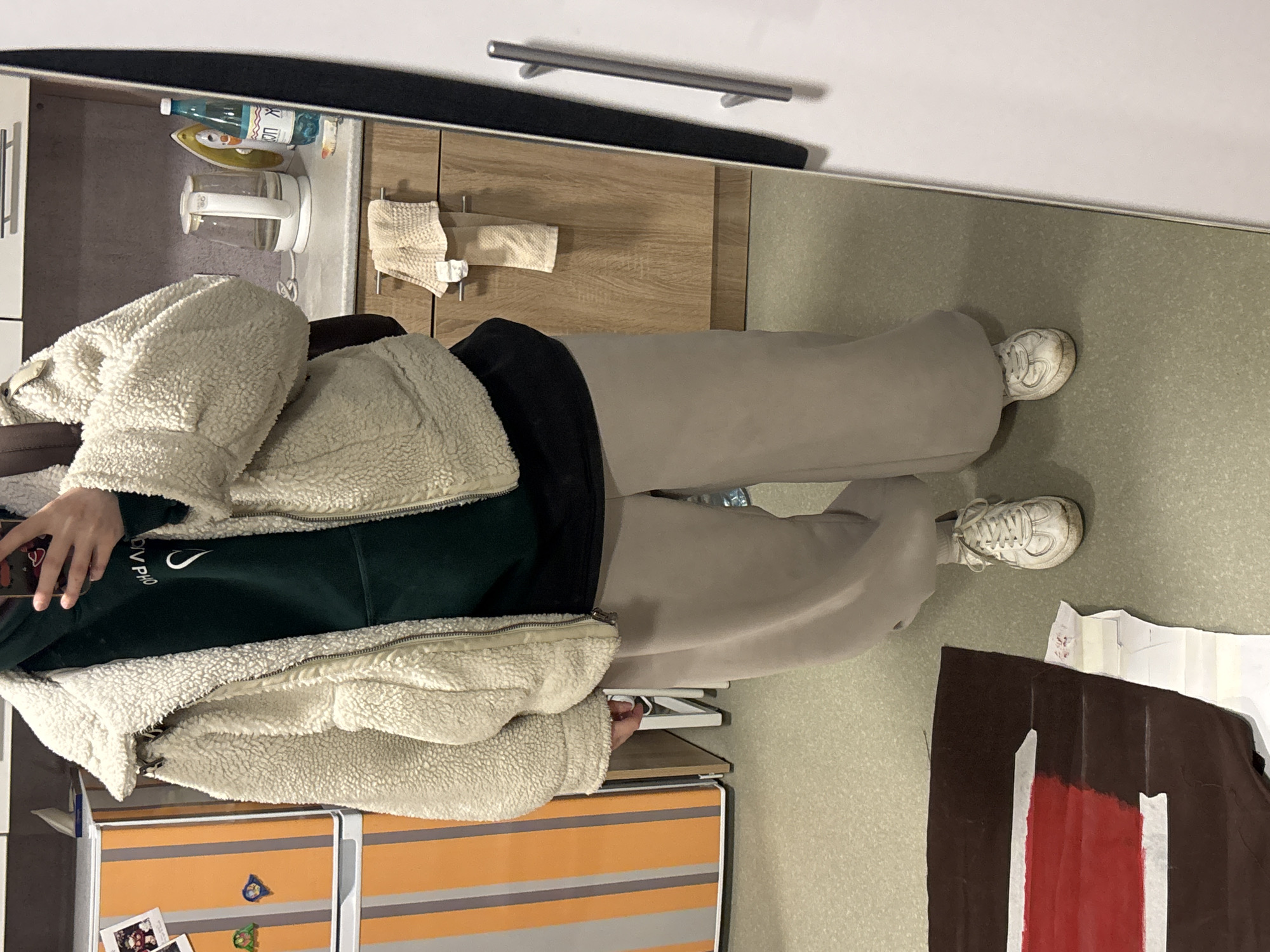 A person wearing a light beige fleece jacket over a dark green sweatshirt and light brown wide-leg pants stands in a room. They are wearing white sneakers. The room appears to be a kitchen with light brown cabinets, a silver handle, and a black appliance on top. A white kettle and other kitchen items are visible on the countertop. On the floor, next to the person, there is a dark brown object with a red stripe. The scene appears to be indoors and could be in an apartment. There is no information in the photo that suggests the location is Perm, Russia.
