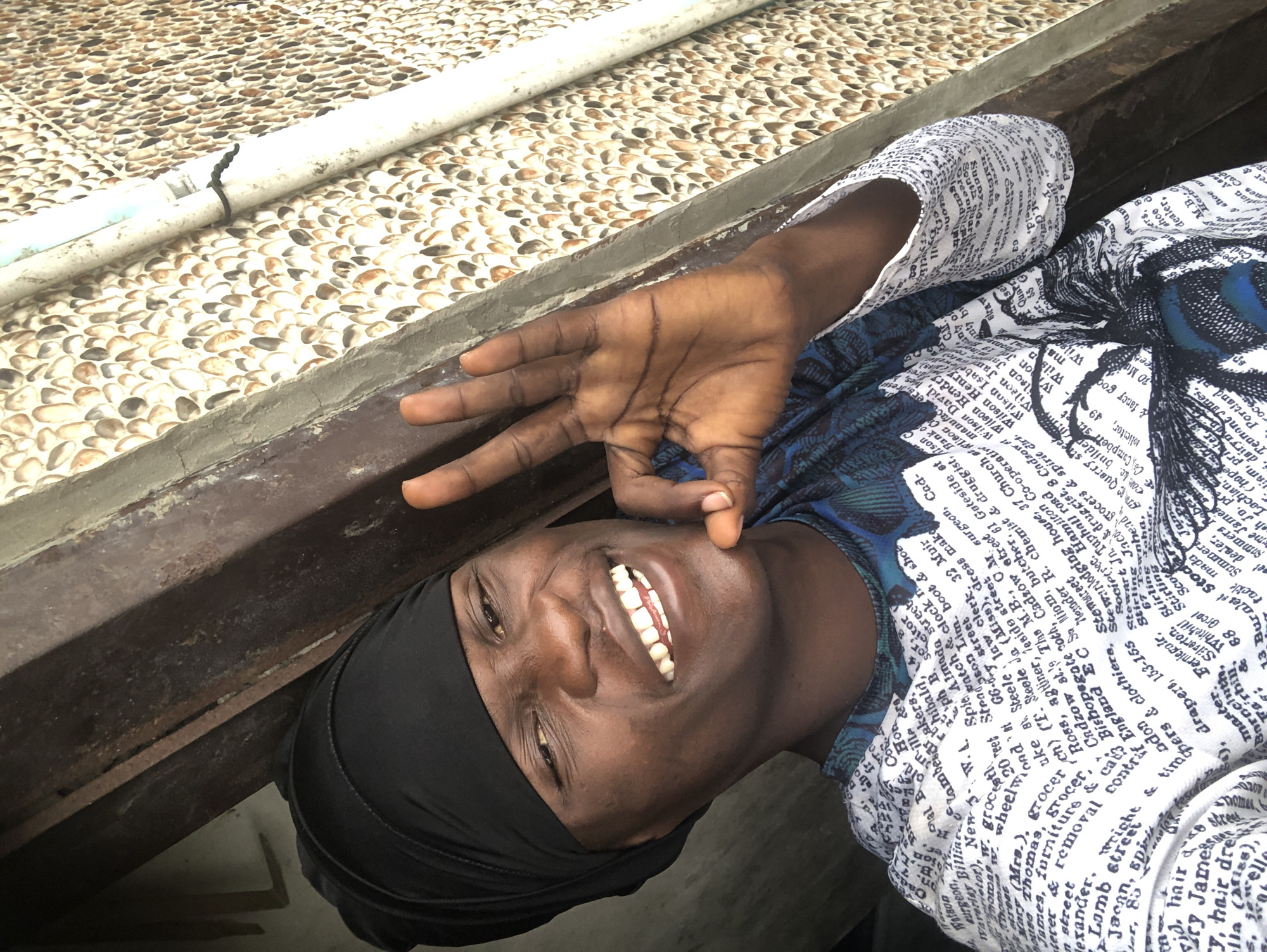 A young Black person, wearing a black head covering and a white shirt with text printed on it, smiles for the camera while making an 'OK' sign with their right hand. They are positioned in the foreground, with a rustic, textured wall featuring small, smooth pebbles serving as the background to the right. A wooden frame or beam is visible on the left, suggesting an outdoor or semi-outdoor setting. The lighting is bright, casting shadows and highlighting the subject's features. The environment suggests a location in Ado, Nigeria, characterized by the distinctive pebble-textured wall, a common architectural feature in some parts of the city.