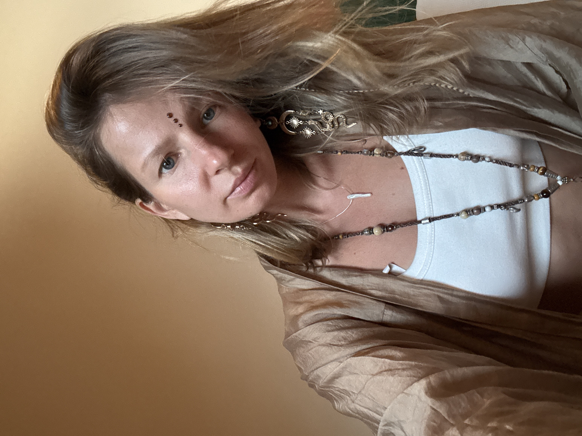 The image is a close-up, tilted selfie of a woman with blonde hair, fair skin, and blue eyes. She is wearing a white tank top and a flowing, shimmering tan-colored garment. Her hair is long and appears slightly windswept. She has a decorative bindi with three small brown stones on her forehead, and elaborate, golden earrings shaped like snakes and moons with stars. A beaded necklace, adorned with various stones and shells, is visible around her neck. The background is a soft, yellowish-brown, suggesting an indoor setting or a backdrop that is out of focus. The lighting appears soft and natural, possibly from natural light sources. The overall mood is serene and artistic, with the woman's gaze directed towards the camera. The tilt of the image and her relaxed expression contribute to an intimate and personal feel. There are no visible signs of text within the image.