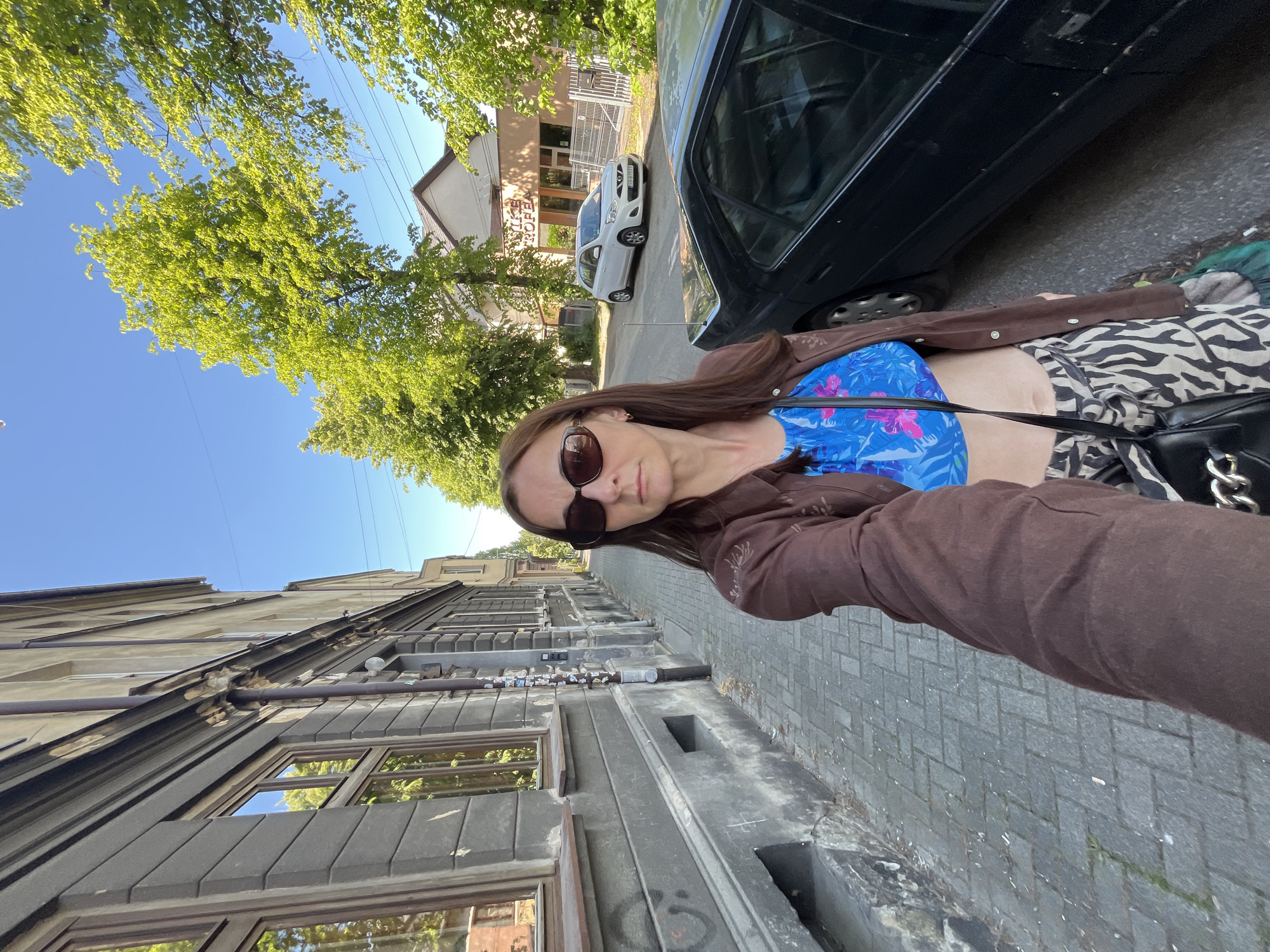 A medium-angle selfie shot captures a woman standing on a paved sidewalk on a sunny day in an unknown city. The woman, with long brown hair, wears dark oval-shaped sunglasses, a bright blue floral crop top, a brown button-up shirt or jacket worn open, and black and white zebra-print pants. A black shoulder bag with silver chain details hangs across her body. Her right arm is extended, holding the camera, and her left arm is mostly obscured.
To her left, a weathered, multi-story building with a grey, possibly stone or stucco, facade runs parallel to the sidewalk. It features recessed windows with brown frames, reflecting green trees and blue sky. A dark vertical drainpipe runs down the building, adorned with numerous small stickers. The sidewalk is paved with grey rectangular blocks in a herringbone pattern.
To her right, an asphalt street is visible, with a dark-colored sedan (possibly black) parked alongside the curb, partially obscured by the woman. Further down the street, a white Nissan Micra (license plate "CD-31 FYY") is parked. Beyond the street, a light-colored building with a brown roof and a sign, partially reading "NIKE TROFEA," stands behind a metal fence. Numerous mature trees with vibrant green foliage line the street, casting dappled light, and their leaves are reflected in the building windows. The sky above is clear and bright blue. Overhead utility lines are faintly visible against the sky.