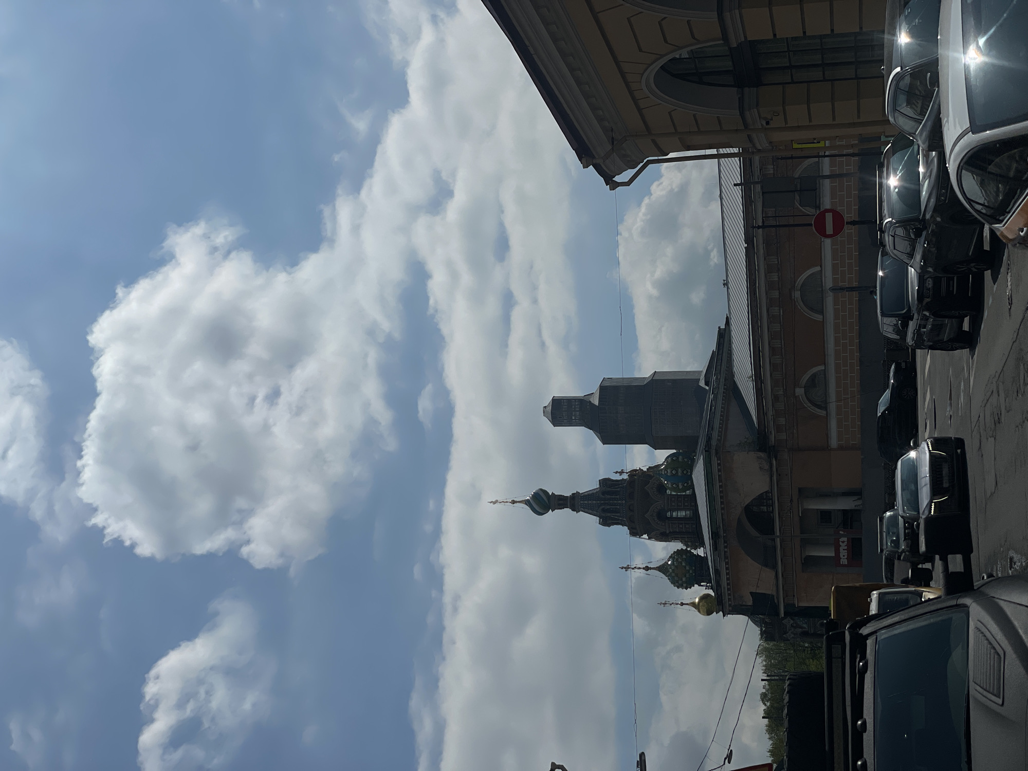 The image shows a street scene in Saint Petersburg, Russia, with the Church of the Savior on Spilled Blood visible. The church's domes and architectural details are prominent against a backdrop of a blue sky with white clouds. Buildings line the street, with cars parked alongside. A 'no entry' road sign is visible among the cars. The image is taken from a low angle, looking upwards toward the church.
