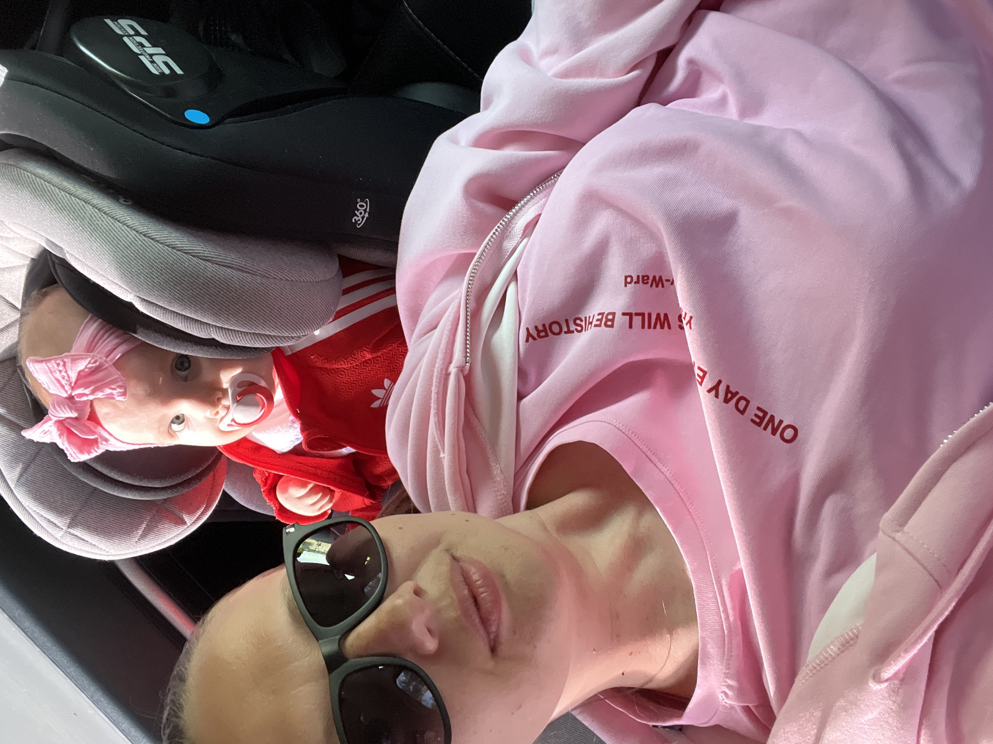 The image, rotated 90 degrees clockwise, displays an adult woman and a baby sharing a car interior, likely during a daytime journey. The woman occupies the lower half of the frame, angled upwards towards the camera. She wears large black sunglasses, obscuring her eyes, and has light-colored hair pulled back. Her lips are slightly parted. She is dressed in a light pink hooded sweatshirt with a zipper, over a white top. Red text is partially visible on her sweatshirt, reading in part "ONE DAY E...", "WILL BE HISTORY", and "-Ward".
Above and behind the woman, a baby is secured in a car seat. The baby has light hair, wide, curious eyes, and a pink bow headband. A pink and white pacifier is in the baby's mouth. The baby is wearing a red long-sleeved outfit, with a partial Adidas logo (three stripes) visible on the shoulder.
The car seat is predominantly black with gray fabric padding. Visible text on the car seat includes "SPS SIDE PROTECTION SYSTEM", "360°", and a small blue circular indicator or button. The strong, direct light suggests a sunny day, consistent with the woman wearing sunglasses. The scene depicts a candid moment, possibly a selfie, within a vehicle. While the image itself doesn't provide external location cues, the context provided states the location is Chaikovsky, Russia.