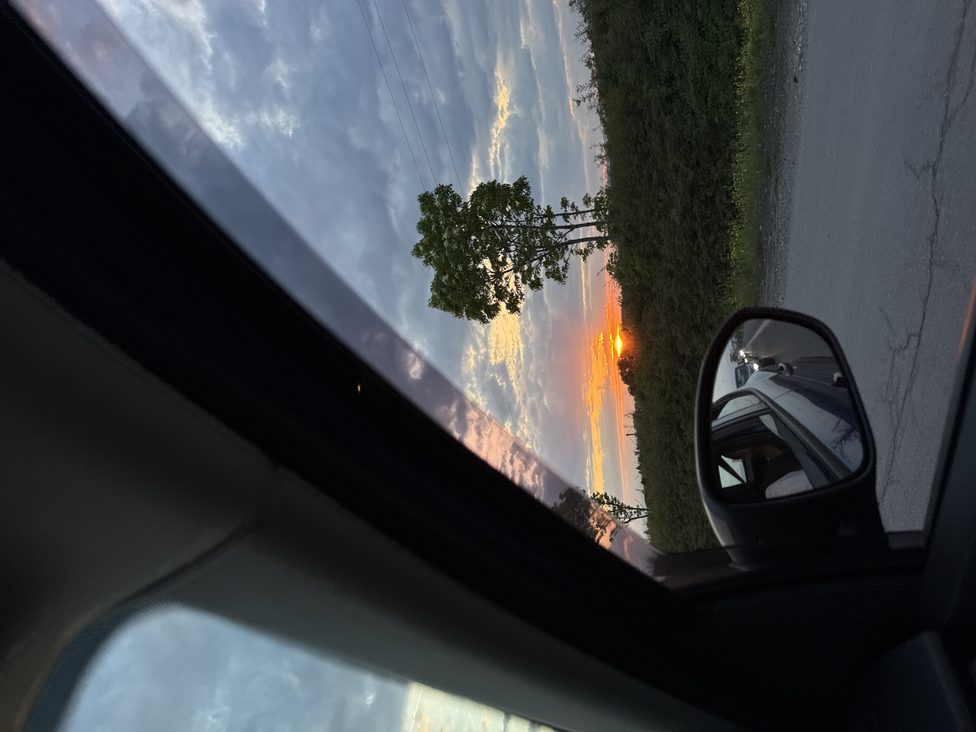 The image depicts a view from inside a car, looking through a sunroof towards a sunset scene. A roadside with green foliage and a gray asphalt road is visible. The setting sun is obscured by foliage and trees. In the side mirror of the car, reflections of other vehicles are apparent. The sky has a blend of blue, white, and orange colors, indicative of the sunset. The scene is not identifiable as Yana, Bulgaria; location cannot be confirmed from visual information.