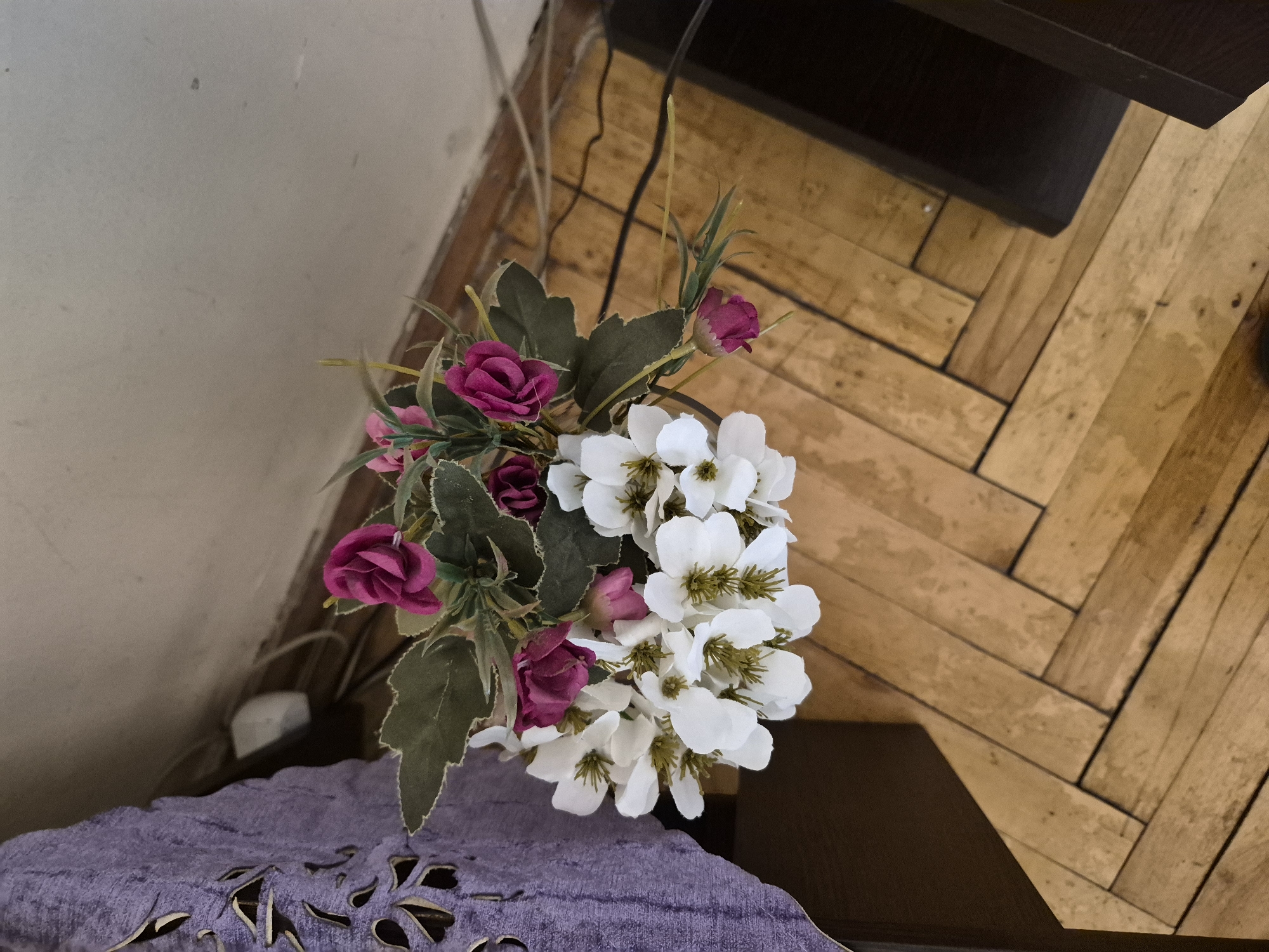 An overhead, slightly angled shot captures a bouquet of artificial flowers alongside elements of an indoor setting in Yerevan, Armenia. The central focus is a cluster of white and magenta (or deep pink) artificial flowers with green leaves, arranged in what appears to be a vase or stand that is not fully visible.
In the foreground, to the bottom-left, a purple fabric object with cut-out or crocheted patterns rests on a dark brown surface, possibly a table or shelf. The fabric shows texture and intricate detailing.
The background primarily consists of a light-colored wall on the left, displaying minor scuffs or marks. To the right and extending across the upper portion of the frame is a wooden parquet floor, laid in a herringbone pattern. The floorboards are light brown with visible grain and some signs of wear or age. Dark electrical wires are noticeable, running vertically along the wall and then horizontally across the wooden floor. A dark brown piece of furniture, likely a shelf or table edge, is visible in the top-right corner and along the right side of the frame, casting a soft shadow on the floor.
The lighting appears to be ambient indoor light, and there are no people or discernible activities taking place. No text is visible in the image.