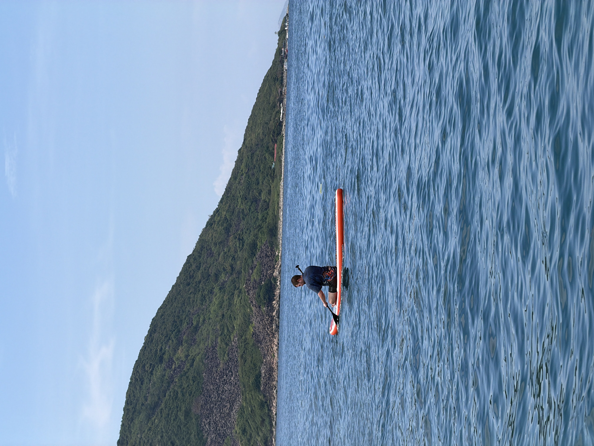 The image depicts a person paddling an orange stand-up paddleboard on the ocean. The paddleboarder is positioned in the water near a coastline. The scene is situated in Bac Nha Trang Ward, Vietnam. The paddleboarder is using a paddle to move through the water. The water surface is rippled by wind. Behind the water and the paddleboarder, a steep, vegetated hillside rises along the coast. A sliver of blue sky is visible above the hillside.