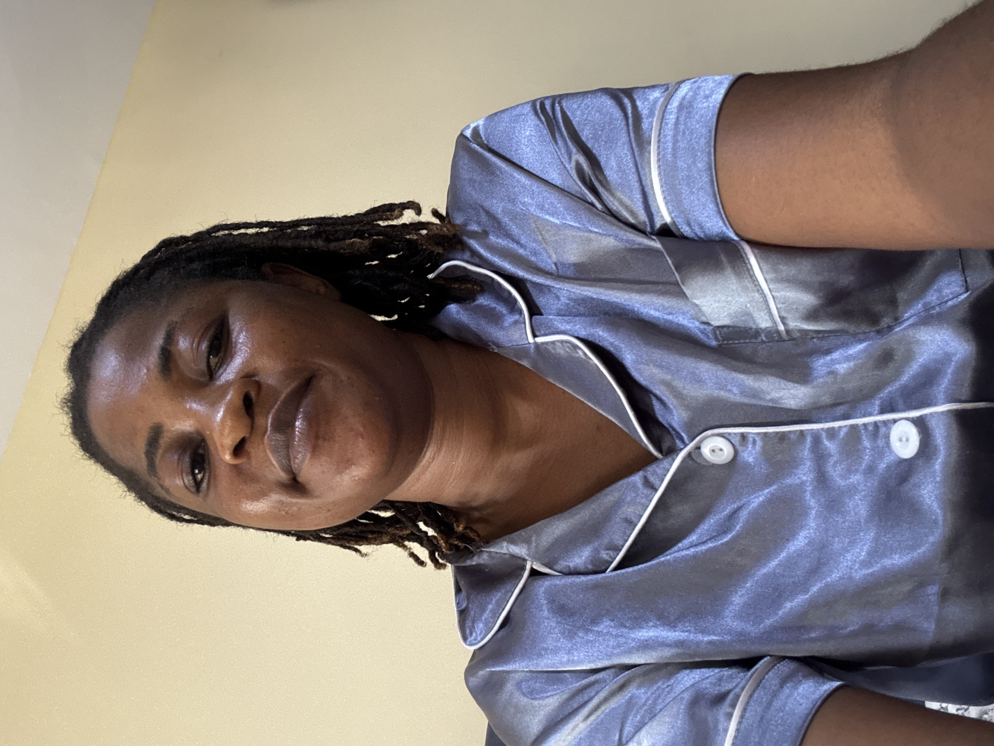 A woman with dreadlocks is looking at the camera. She is wearing a blue satin pajama top with white piping and buttons. The background is a plain cream-colored wall. The image appears to be a selfie taken indoors. The lighting is soft and even. There are no visible indications of the specific location within Ajara, Nigeria.