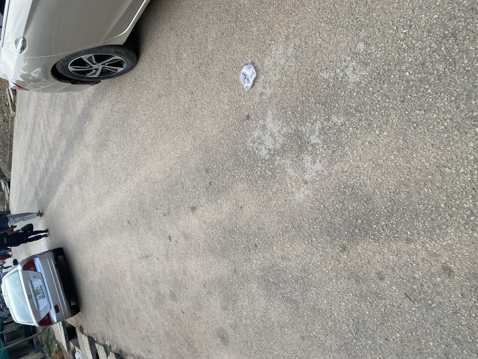 An outdoor, eye-level, medium shot captures a street scene in Jos, Nigeria. The foreground is dominated by a grey asphalt road surface. To the left, a silver Honda Civic sedan is parked, its rear end visible with the license plate "RBC-855EH". Two individuals, a man and a woman, walk away from the viewer on the right side of the road. The man is wearing black trousers and a black shirt and is carrying a black bag. The woman is wearing light-colored trousers and a dark top. In the middle ground of the road, there is a single crumpled piece of paper lying on the asphalt. The background features an unpaved area with scattered vegetation and some concrete structures, suggesting a semi-urban or undeveloped area within the city. The lighting indicates it is daytime.