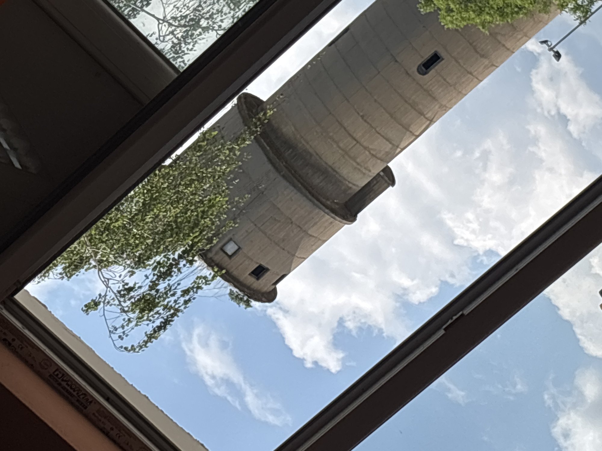 The image captures an upward view through a window or skylight, framing a tall, cylindrical concrete structure against a bright blue sky with scattered white clouds. The structure, likely a water tower or silo, is made of light grey concrete with visible horizontal segment lines. Several small, dark, rectangular openings resembling windows are visible on its surface. Green foliage and tree branches are seen on the left side, with some vines or growth clinging to parts of the concrete tower.
On the upper right, beyond the main structure, a utility pole with two dark light fixtures is visible against the sky. The dark frames of the window or skylight partially obscure the top, bottom, and right edges of the scene. On the bottom left window frame, faint white text, including "PROFIN" and "CE", is visible. The scene suggests a clear, sunny daytime environment. No people or specific activities are depicted; it is a static architectural and natural landscape view.