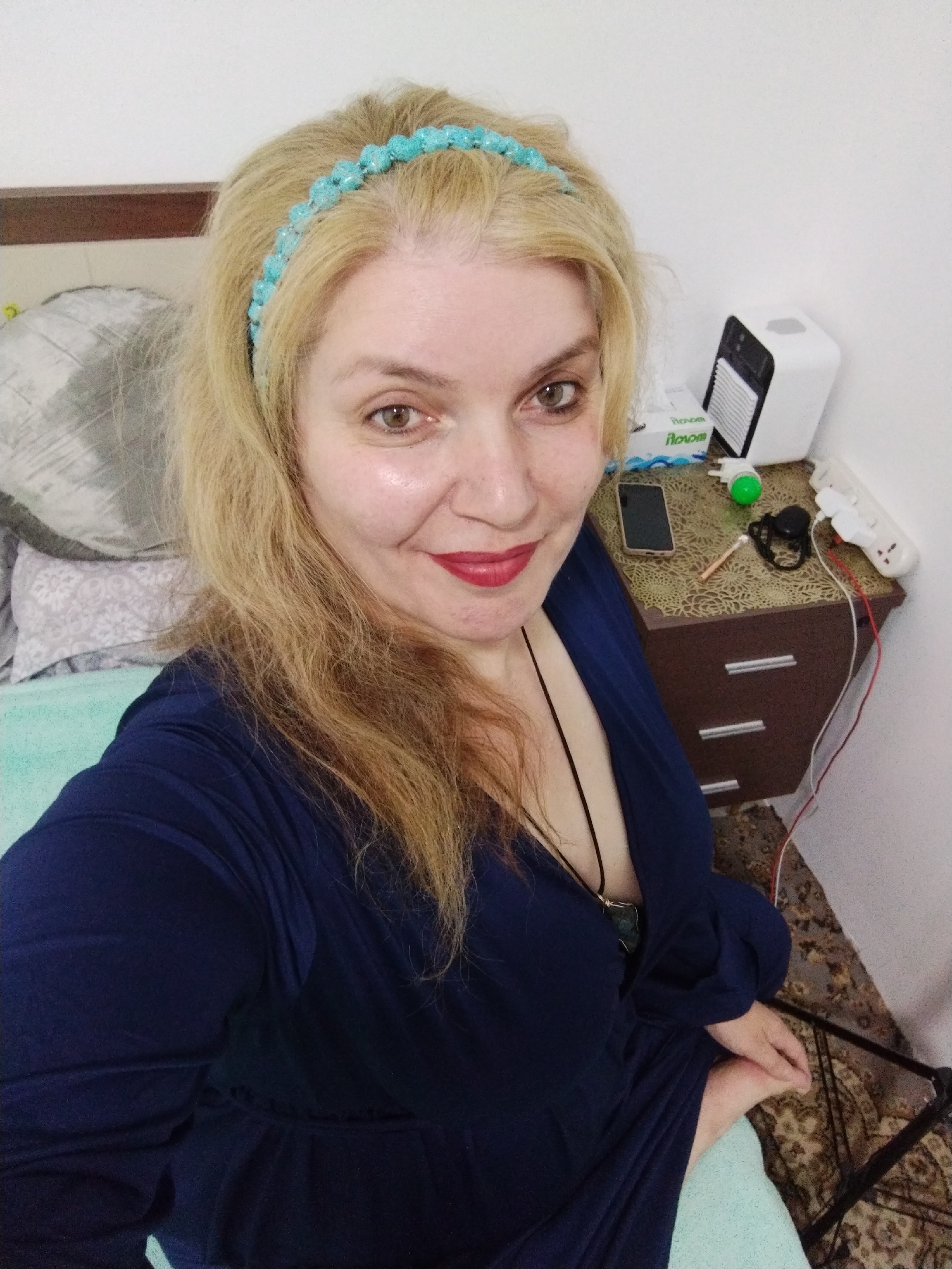 The image depicts a woman with blonde hair and a turquoise headband. She is wearing a dark blue dress and red lipstick. The background shows a bed with a patterned pillow and a mint green sheet. A bedside table holds a cell phone, a small white air conditioner, a tissue box, and an electrical outlet strip with various items plugged in. The location is likely a bedroom, potentially in a residential setting. There is no definitive information to confirm this is the city of Al Mas'oudi, United Arab Emirates.