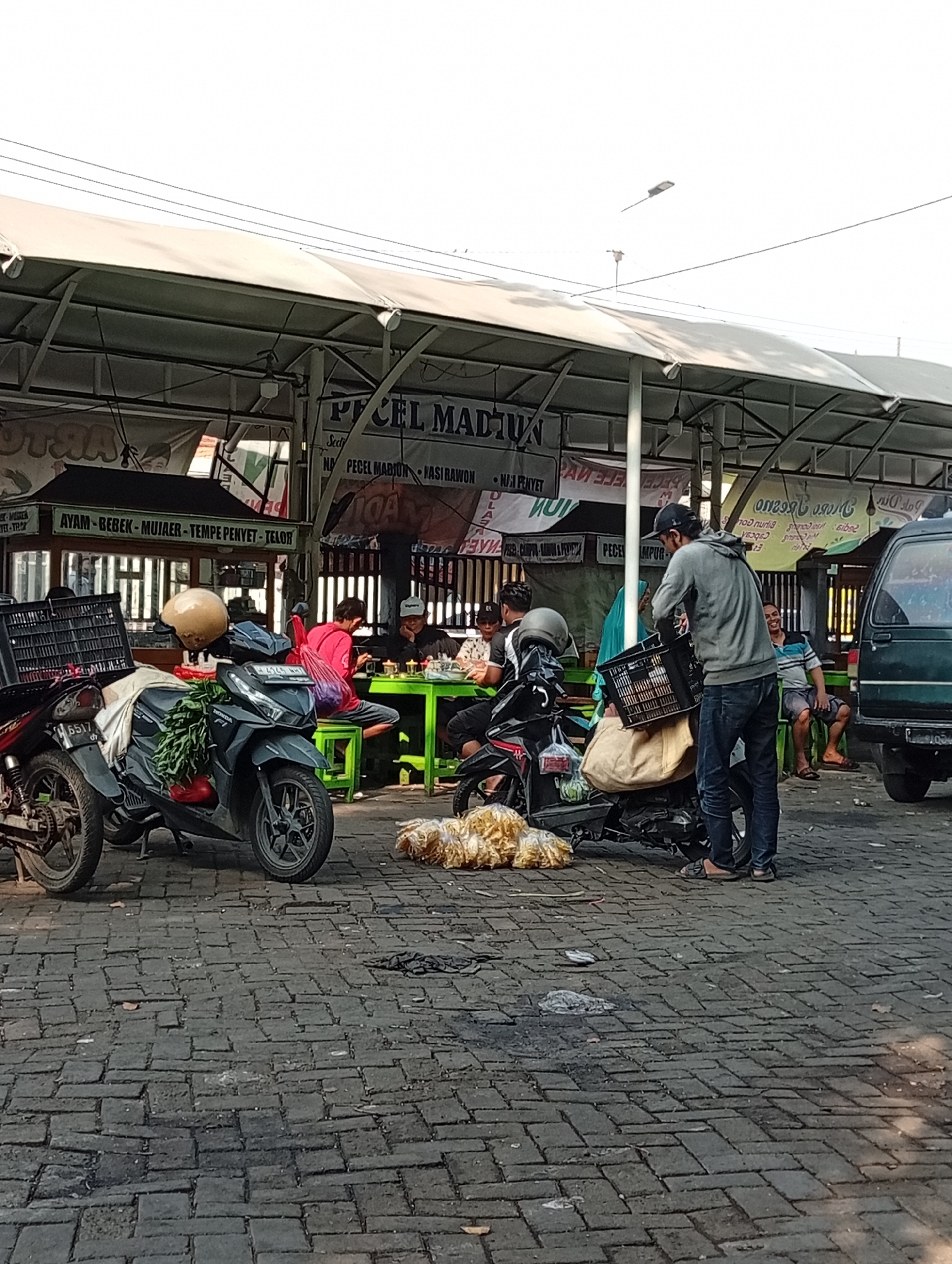 This is a street-level scene in Larangan, Indonesia, capturing a bustling food stall area under an awning. The foreground is paved with dark cobblestones, showing signs of wear and tear. Two motorcycles are parked prominently, one in the left foreground with a black crate on its back and a pile of green vegetables tied to its handlebars. The other motorcycle, a dark gray scooter, is in the center, with a yellow helmet on its seat and a large sack of what appears to be dried goods or snacks behind it.
In the mid-ground, several people are seated at green tables under the awning, eating. One man in a red shirt is particularly visible, leaning forward. Further back, a man in a gray hoodie and jeans is carrying a black crate, appearing to be a vendor or customer interacting with the stall. Another motorcycle is parked behind him. A green van is visible on the far right of the frame, parked near the stall area. Signs above the stalls indicate food offerings, with "PECEL MADUN" being a prominent name. The overall impression is of a vibrant, everyday street food scene in an Indonesian city.