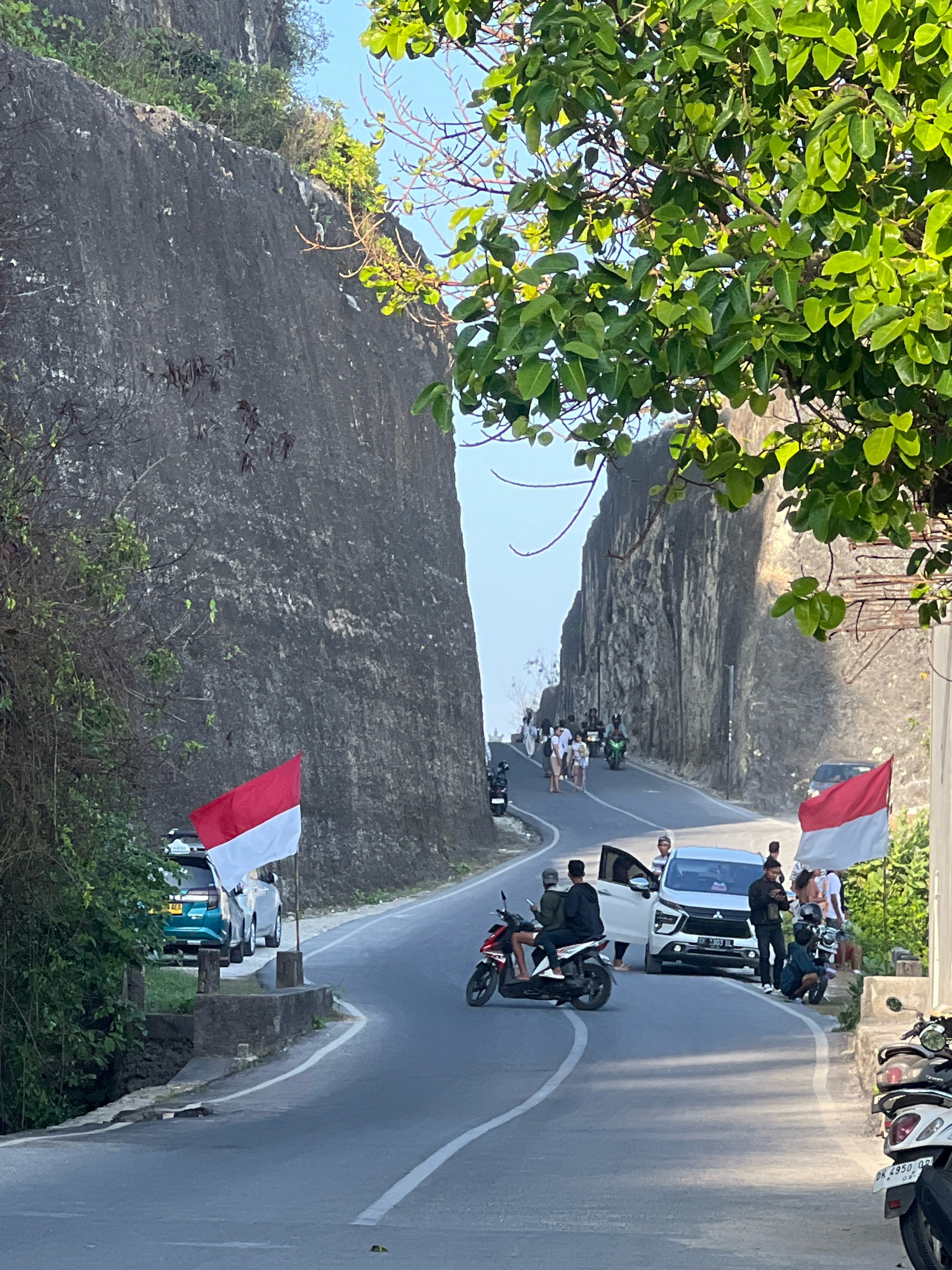 A road curves between steep, grey, rocky cliffs in Kuta Selatan, Indonesia. Lush green trees with vibrant leaves overhang the scene from the top right. Two Indonesian flags, red and white, are prominently displayed along the roadside, one near a parked teal car and the other further down the road. Several people are visible, some standing and observing, while others are on motorcycles and in cars. A white SUV with its door open is stopped, and a group of people are gathered around it. A man is kneeling next to it, looking at his phone. On the road, two men on a scooter are heading away from the viewer, and another scooter is parked on the left. Several parked motorcycles are also visible on the right side of the road. The sky is a clear, light blue. The road is paved and has white lane markings.