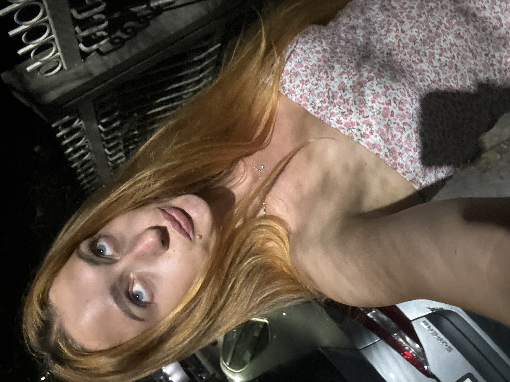 A close-up, low-angle selfie shows a young woman with long, reddish-blonde hair. She is wearing a pink floral dress. Her head is tilted back and to the side, with her eyes wide and looking upwards. She has makeup around her eyes and her lips are slightly parted. A thin silver necklace with a small pendant is visible around her neck.
In the background, a black metal fence with decorative circular elements is visible on the left. Part of a silver car, including a taillight and part of the bumper, is visible on the right. The lighting suggests it is nighttime, with a shadow cast by what appears to be a phone, indicating the selfie was taken with a handheld device. The overall scene appears to be outdoors, possibly in a residential area or near a building with a decorative fence. The woman's expression could be interpreted as surprised or questioning.
