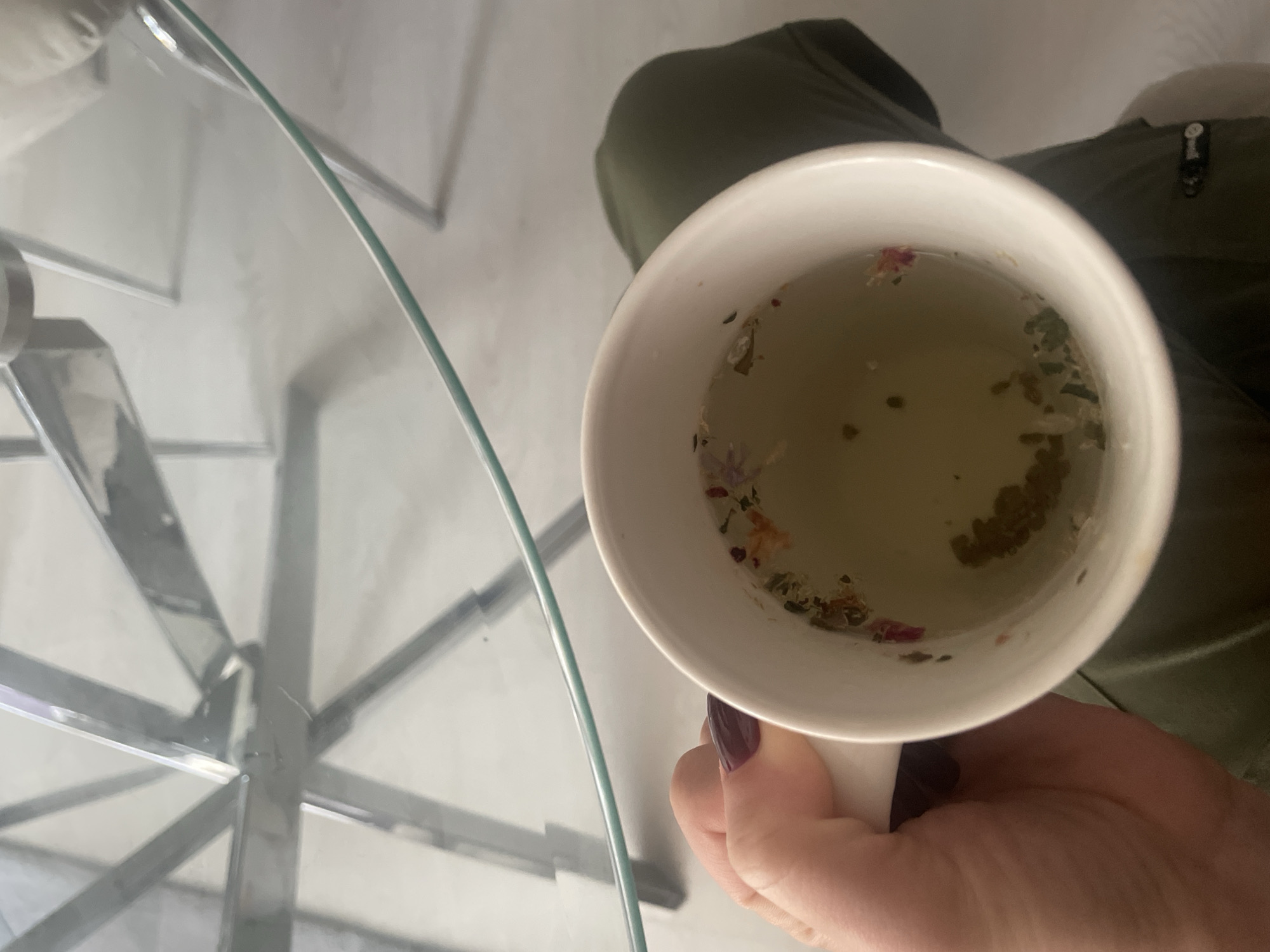 A white ceramic mug filled with light brown liquid containing visible plant matter, including small flower petals, is held by a hand with dark purple nail polish. The mug is resting on a clear glass table with a visible metal frame. In the background, there is a green garment with a zipper visible. The location is unknown and there is nothing in the image to suggest that it is the city of Sofia, Bulgaria.