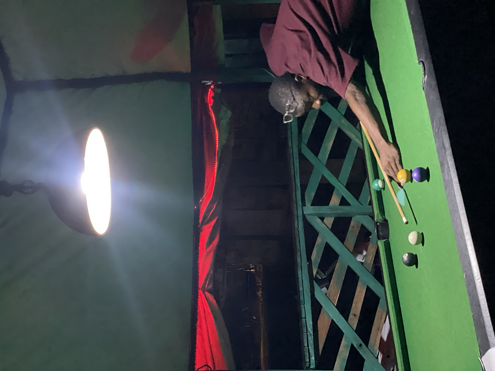 In Lokoja, Nigeria, a man with glasses wearing a maroon shirt is playing pool. He is positioned at a green pool table, leaning forward to strike a ball with a cue stick. Several billiard balls are scattered on the table, including a black 8-ball, a white ball, a yellow ball, and a purple ball. The setting appears to be outdoors, possibly a bar or entertainment area, with a green wooden railing visible in the foreground. Overhead, a bright lamp illuminates the scene, casting shadows. In the background, red fabric is draped, creating a decorative element, and beyond that, a dark, indistinct space.