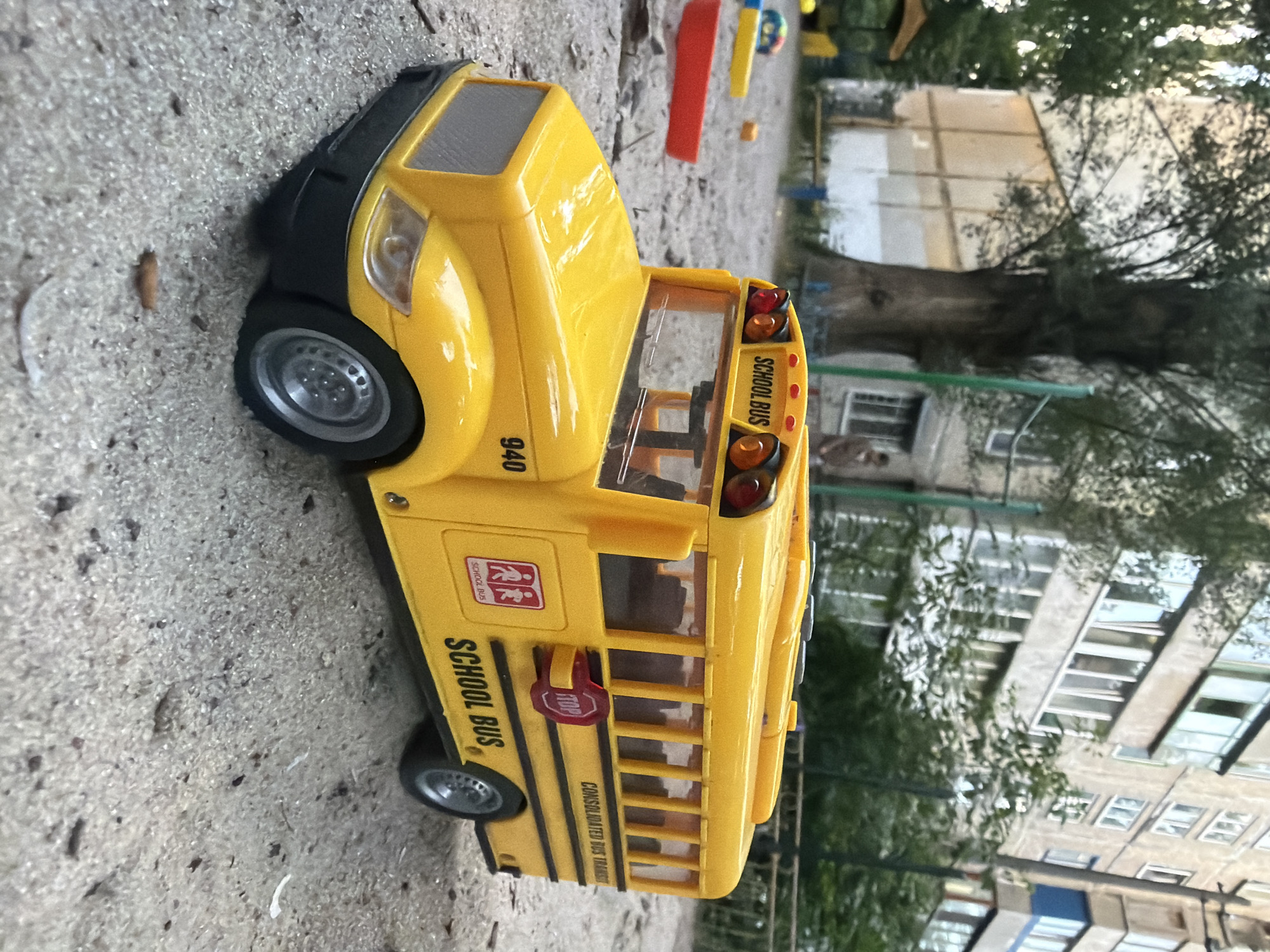 A toy yellow school bus is parked on a sandy playground. The bus has "SCHOOL BUS" written on the back and side. A red stop sign is attached to the side of the bus. In the background, a tall, leafy tree stands in front of a light-colored apartment building. Other playground equipment, including a red plastic toy, is visible on the sand. The scene appears to be in an urban environment, possibly a residential courtyard.