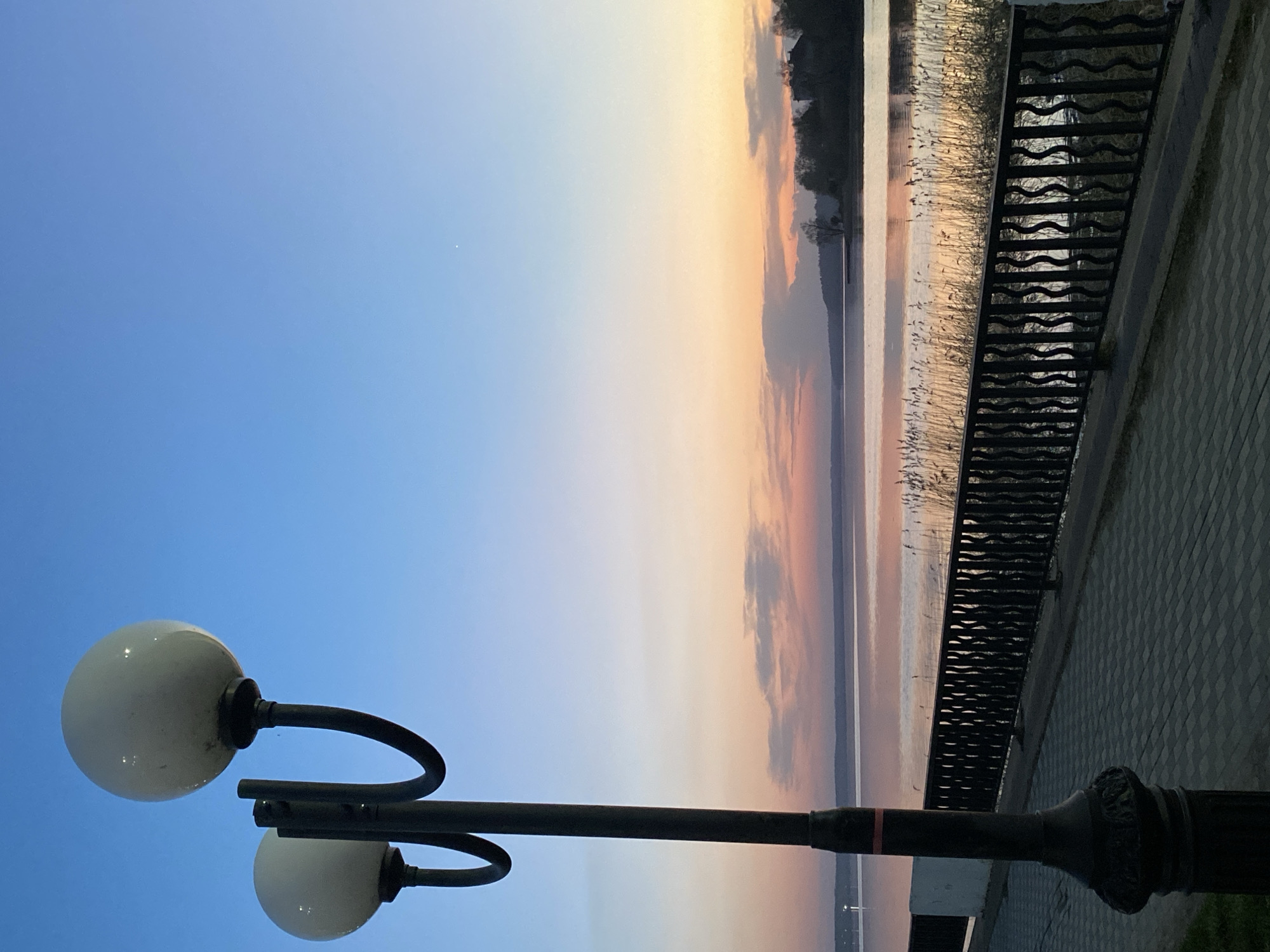 The image shows a street scene in Votkinsk, Russia, at dusk or dawn. The sky transitions from blue to orange and pink hues. A body of water, possibly a lake or river, is visible in the background, reflecting the colors of the sky. A decorative metal railing runs along the edge of a paved walkway. A street lamp with two globe-shaped lights is in the foreground. The lamp post is black with decorative details. The overall scene conveys a calm and serene atmosphere.