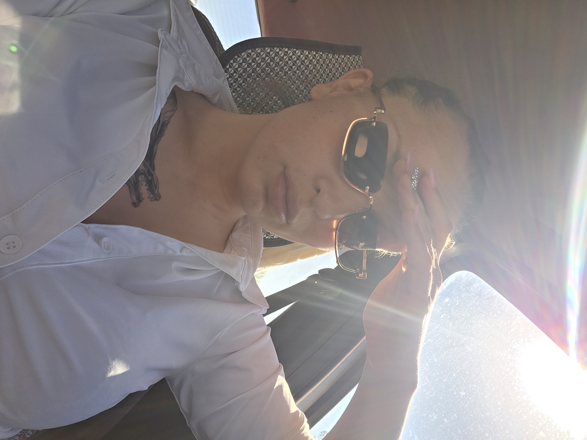 A woman wearing sunglasses and a white button-down shirt is in a car. She is partially obscuring her face with her right hand. A tattoo is visible on her upper chest. The background is bright due to sunlight. The location is Piperevo, Bulgaria.