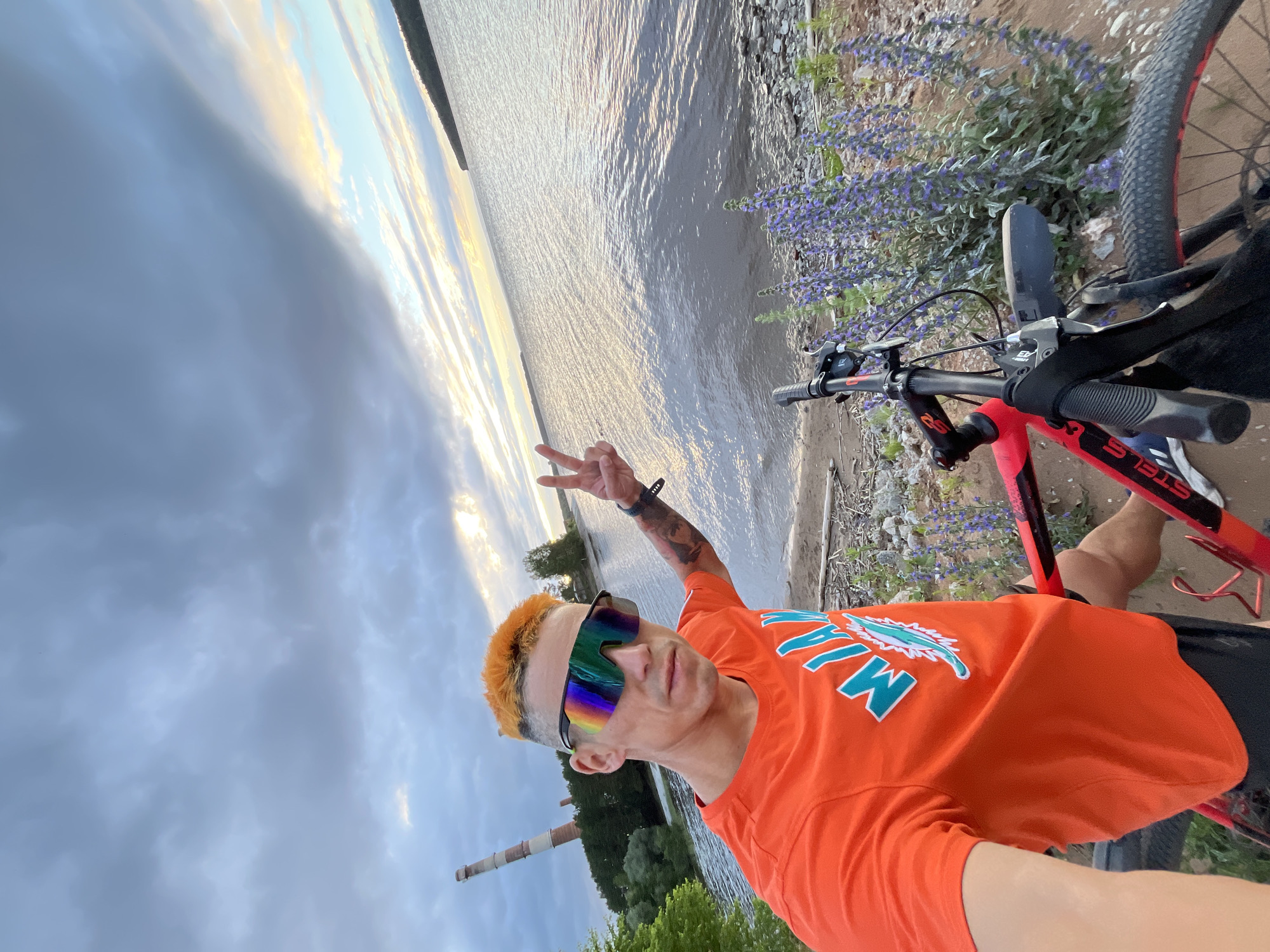A person with bright orange hair and mirrored sunglasses is taking a selfie outdoors. They are wearing an orange t-shirt with a turquoise logo that appears to be a dolphin. Their left hand is raised in a peace sign. A red and black bicycle is parked next to them. The scene is by a body of water, possibly a river or lake, with a shoreline covered in pebbles and purple flowers. In the background, there is a tall, cylindrical industrial structure with a lighter color and a darker band near the top, which is likely a smokestack. The sky is cloudy with patches of sunlight breaking through, suggesting it might be late afternoon or early evening. The overall mood appears casual and relaxed, with the person enjoying a moment by the water with their bicycle. The location context is Chaikovsky, Russia.