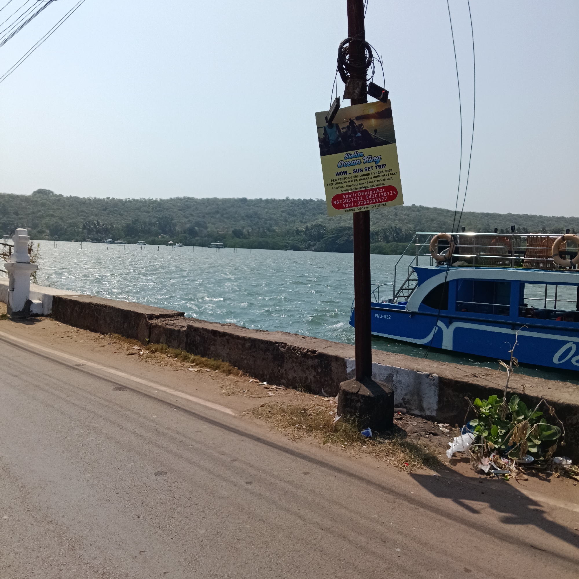 The photograph depicts a waterside scene in Oxel, India. A paved road runs along a low concrete barrier, beyond which lies a body of water with mild waves. In the background, there is a densely vegetated shoreline. A blue and white boat is docked beside the barrier, with the partial visible registration number "PNJ-932". Adjacent to the boat, some foliage grows amongst discarded trash. Attached to a utility pole is a promotional sign for "Siolim Ocean King" sunset trips, with contact information and timing details. The sky is clear and bright.
