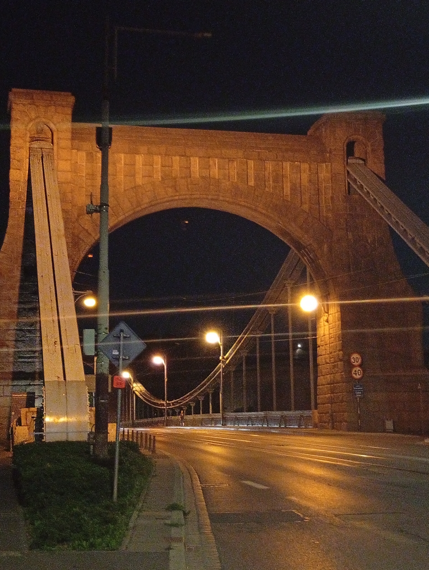 The image depicts the Grunwaldzki Bridge in Wrocław, Poland, at night. The bridge features a prominent archway made of brick and stone. Streetlights illuminate the road, casting long shadows and creating a warm glow on the wet asphalt. Traffic signs, including speed limits of 30 and 40, are visible on the side of the road. The suspension cables of the bridge are also visible. The surrounding area appears to be a pathway with some green shrubbery. The sky is dark, indicating it is nighttime. There are no people or vehicles clearly visible in the frame.