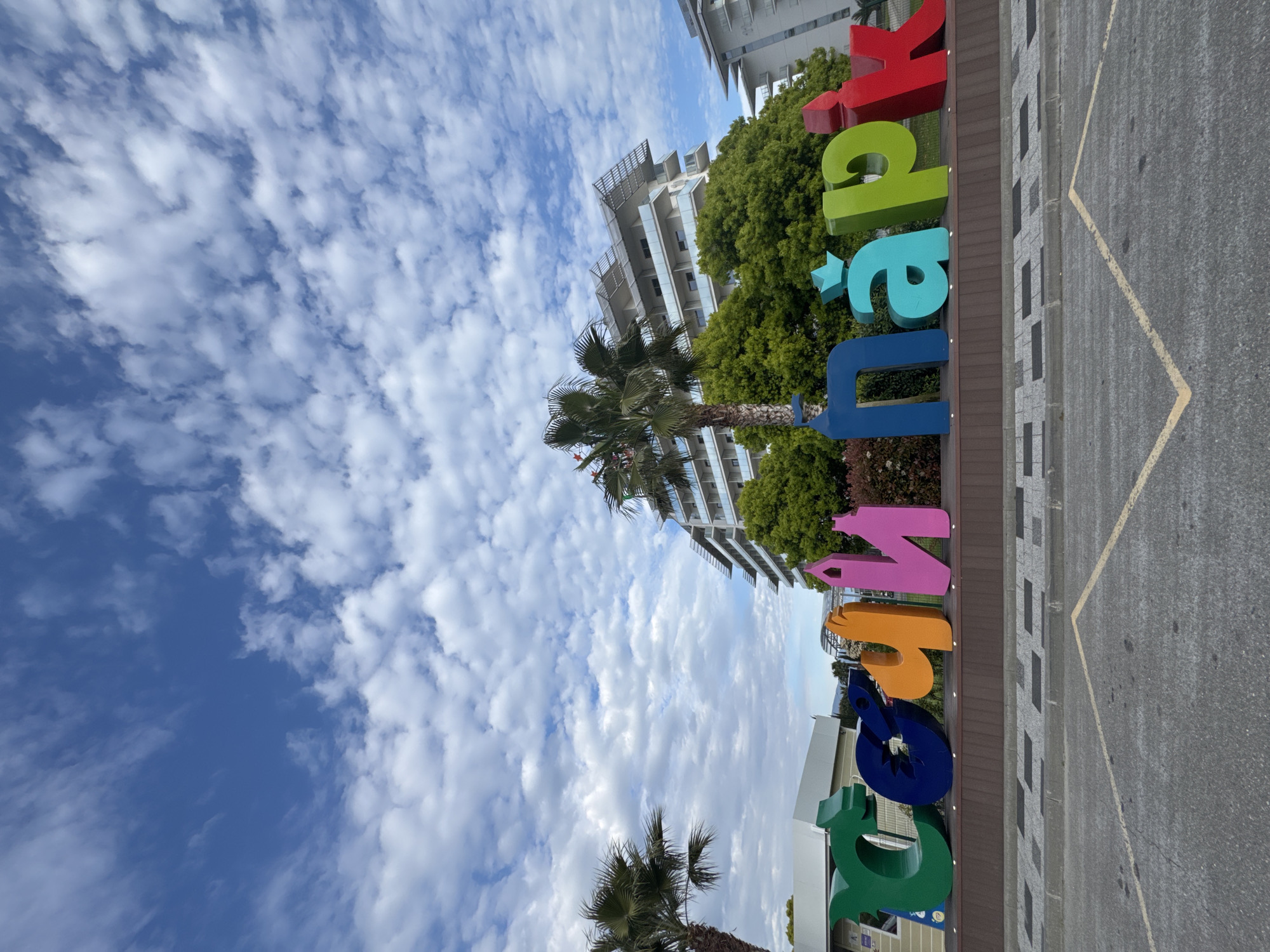 The image shows an outdoor scene with a colorful "Sirius Park" sign in the foreground. The letters are large and each one is a different color (green, blue, orange, pink, blue, teal, green, and red). The sign is situated on a wooden platform. Behind the sign are trees, a palm tree, and a multi-story building with a modern design, featuring balconies and glass elements. The sky is visible, filled with numerous white clouds and patches of blue. A paved area with painted lines (possibly a parking lot) is visible in the right side of the image.