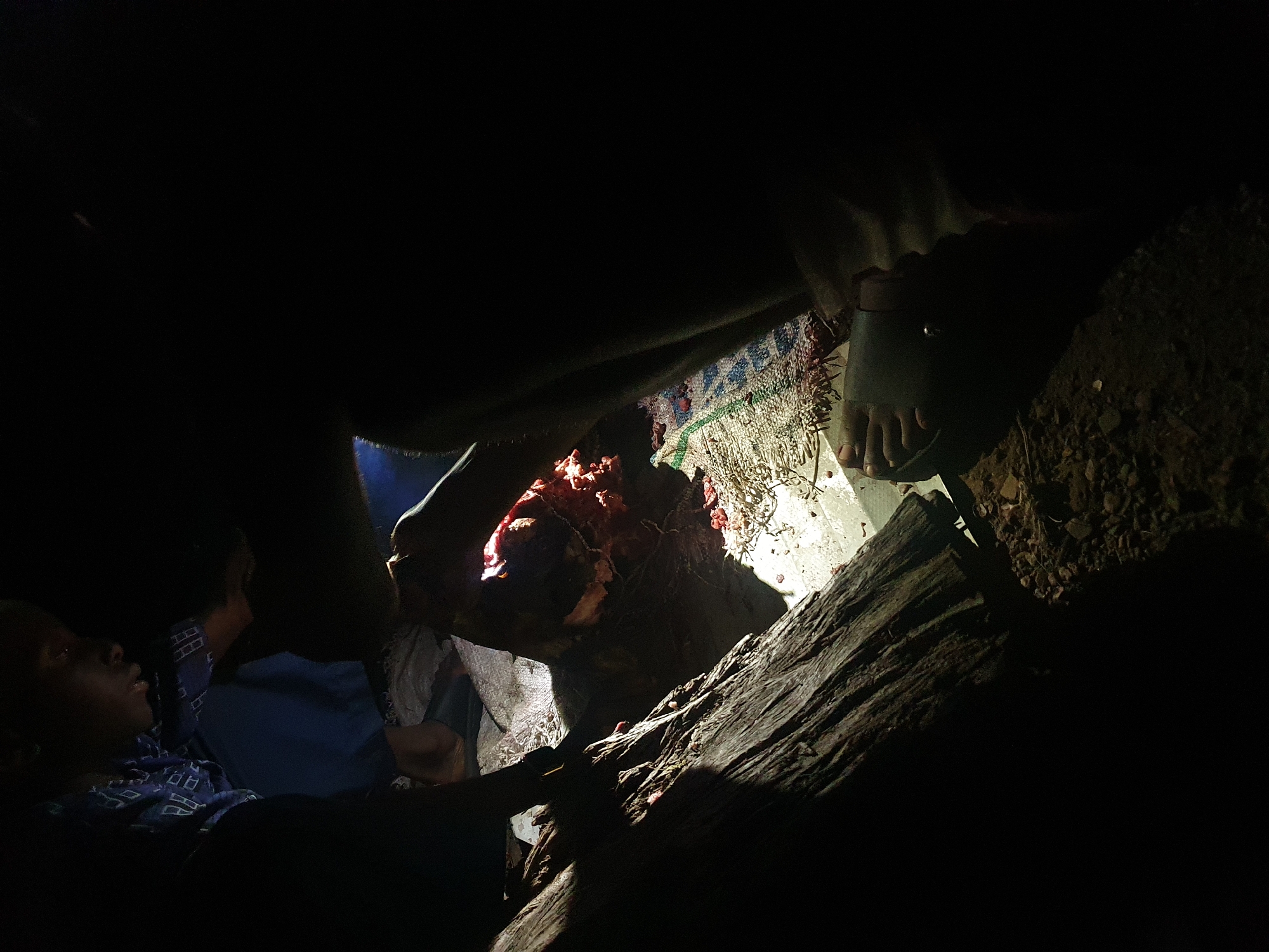 In a dimly lit setting, possibly a confined space in Idah, Nigeria, two individuals are present. One person lies down, visible from the chest up, wearing a patterned blue and black shirt. Another person sits up beside them, wearing a blue shirt. A foot in a sandal is positioned on a textured mat next to a pile of dirt. In the center, a mass of meat rests on a piece of wood or a tree stump.