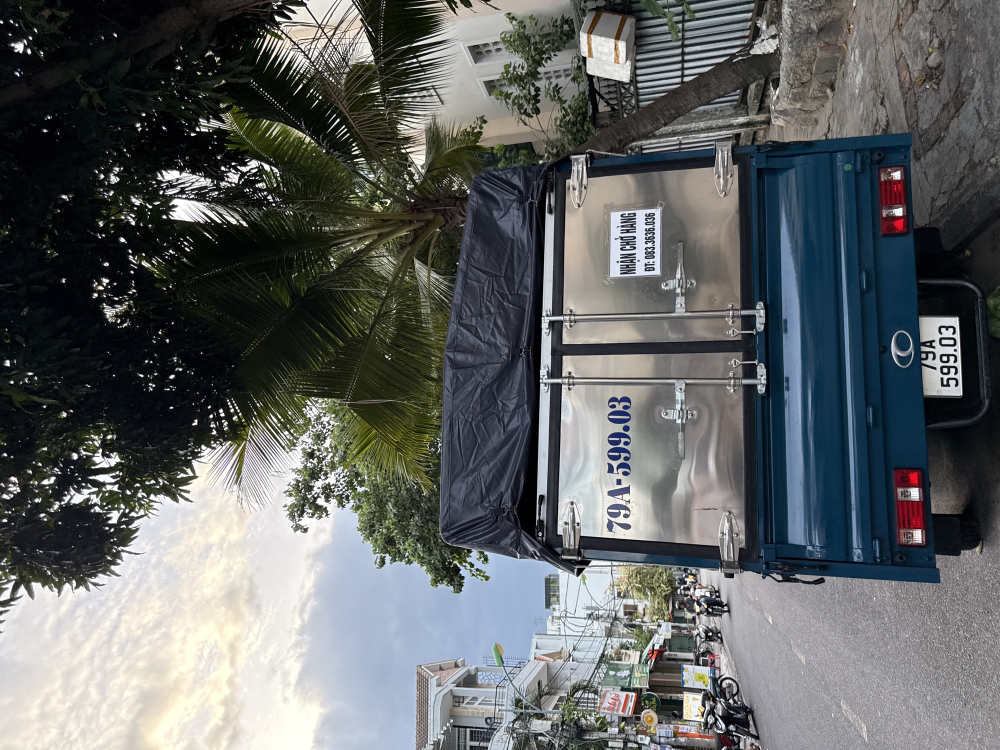 In Nha Trang, Vietnam, a blue truck is parked on the right side of a street. The truck has a stainless steel cargo box covered with a black tarp. The license plate on the back of the truck reads "79A-599.03". On the side of the cargo box, there is a white sticker with Vietnamese text that translates to "Receiving goods" and a phone number "083.3636.036". The street is lined with buildings, trees, and other vehicles, including motorbikes. The sky is cloudy.