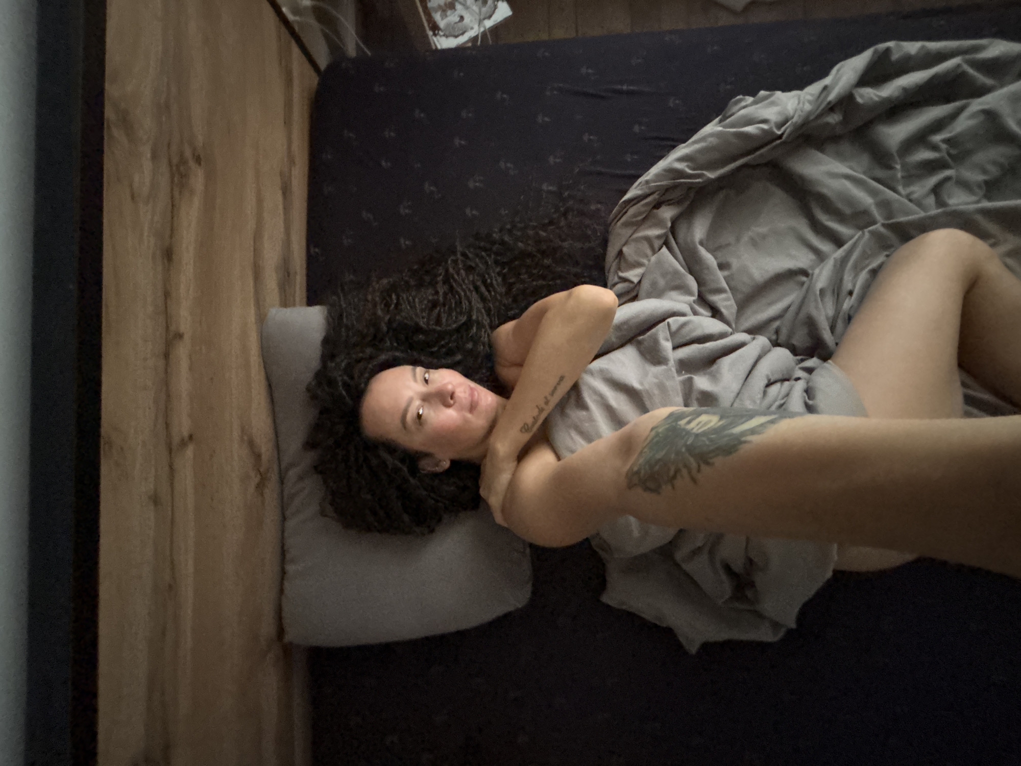 A person with curly hair is lying in bed on a gray pillow. The bed has a dark blue sheet and a gray blanket partially covering the person. The person has tattoos on their arms. A wall is visible on the left side of the frame. The image was taken indoors. The location and city of Chaikovsky, Russia are not discernible from the image.