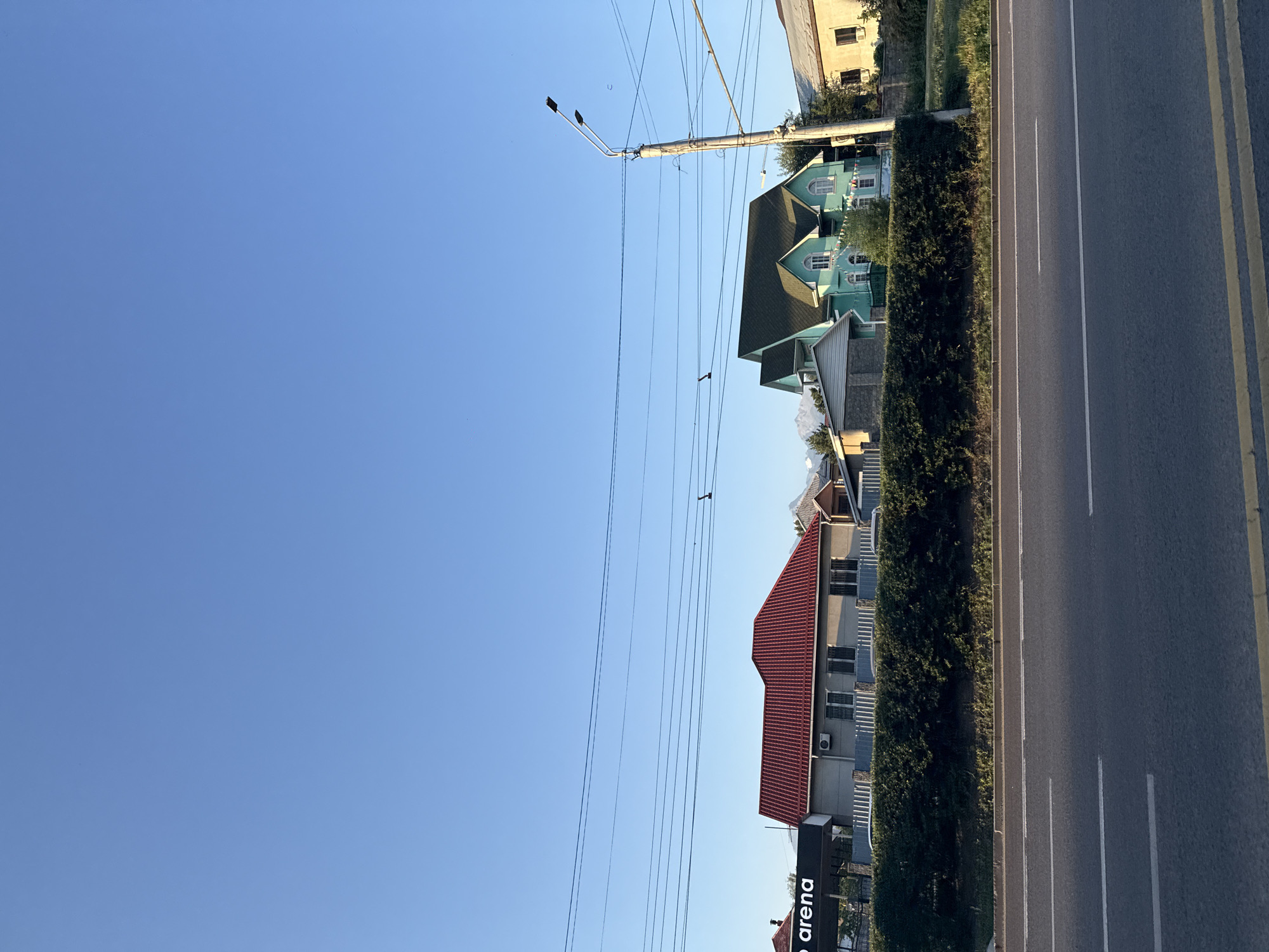 The image captures a street scene in Almaty, Kazakhstan, under a clear blue sky. In the foreground, a two-lane asphalt road with white lane markings is visible. A low, dense hedge runs along the side of the road, separating it from a row of houses and a building with a red roof. To the left of the red-roofed building, a dark structure with the illuminated text "o arena" is partially visible. Further to the right, a vibrant turquoise house with a dark green roof stands out. A wooden utility pole with two streetlights and multiple power lines runs vertically near the turquoise house. The houses appear to be residential, with windows and architectural details suggesting a suburban or semi-urban setting. In the background, distant mountains are faintly visible through the clear sky. The lighting suggests it is daytime, likely late afternoon due to the long shadows cast by the utility pole and the subtle warmth of the light on the buildings. No people are clearly visible in the frame.