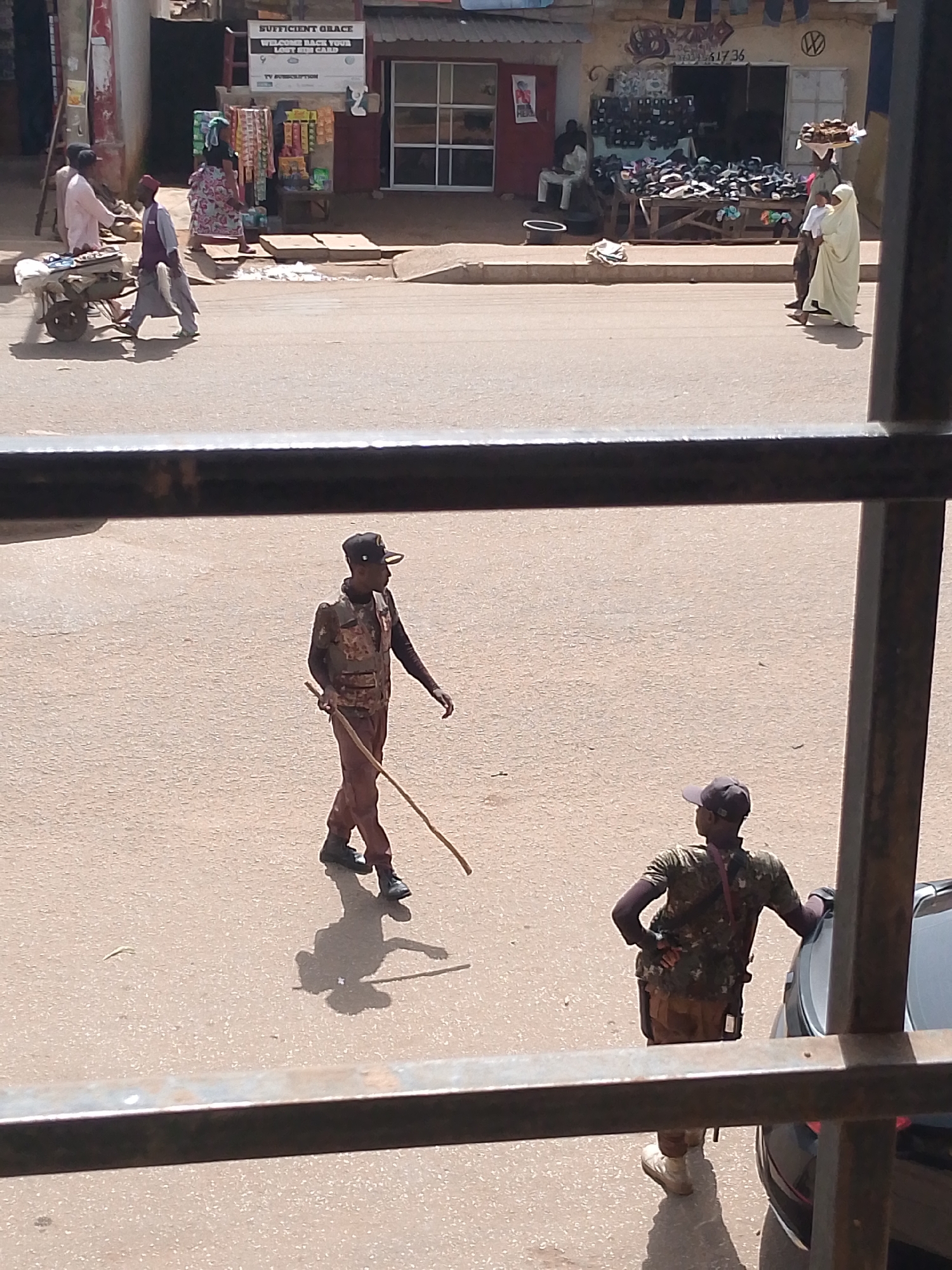 The image depicts a street scene in what appears to be a market area in Bukuru, Nigeria. The foreground shows a dark metal bar, suggesting the photo was taken from behind a window or railing.
In the mid-ground, two individuals dressed in military-style uniforms are visible. One person, facing left and walking, is holding a stick. They are wearing a cap, a camouflage vest over a dark long-sleeved shirt, and brown trousers. Their shadow is cast on the sandy ground. The second person, facing away from the camera and towards a black car, is also wearing a cap, a camouflage shirt, and tan trousers. They have their hands on their hips, leaning slightly against the car.
In the background, several people are walking and interacting near shops. One person in a pink outfit is near a cart with items on it. Another person in a patterned dress is walking past a stall displaying various goods, including packaged items. Further down the street, a person in a light-colored garment is walking, carrying something on their head. The buildings in the background are typical of a marketplace with open storefronts displaying goods like footwear and other merchandise.
The time of day appears to be daytime, with strong shadows indicating sunlight. The weather seems to be warm and dry, consistent with a sunny day in Nigeria. The scene suggests a busy commercial environment. Visible text includes "SUFFICIENT GRACE WELCOME BACK YOUR LAST SIM CARD" on a sign above one of the shops.