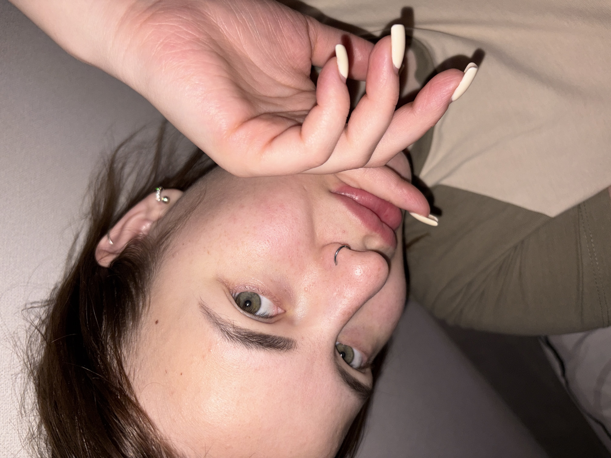 A close-up, upside-down portrait of a young woman with fair skin and light brown hair. She has a nose piercing and earrings. Her left hand is raised, partially covering her face, with long, artificial nails painted a light beige color. The background consists of a beige-colored fabric, possibly clothing or bedding, and a section of a gray pillow. The location is undetermined as there are no contextual indicators within the image.