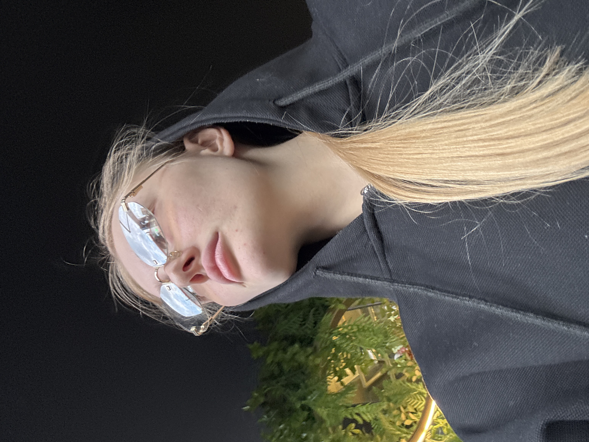 A young woman with light blonde hair and wearing glasses is looking towards the bottom left of the frame. She is wearing a dark hoodie. The background is dark, but a bush is visible near the bottom of the frame. The exact location in Perm, Russia is not discernible from the image.