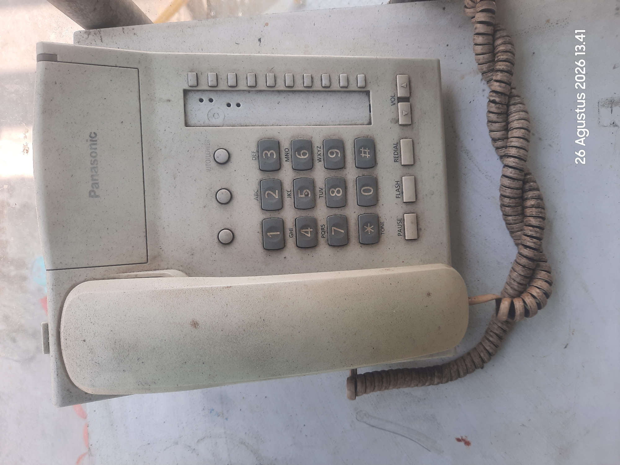 The image shows a dusty, beige Panasonic corded telephone placed on a light-colored, textured surface, likely outdoors. The telephone handset is resting in its cradle, and the coiled cord is visible. The keypad is standard, with numbers and symbols, and there are several buttons for functions like PAUSE, FLASH, REDIAL, and VOLUME control. The brand "Panasonic" is printed on the top of the telephone. The background is blurred, but appears to be a wall or surface with some smudges and debris. A timestamp in the lower right corner reads "26 Agustus 2026 13.41". The image does not provide specific details about the location within Gending, Indonesia.