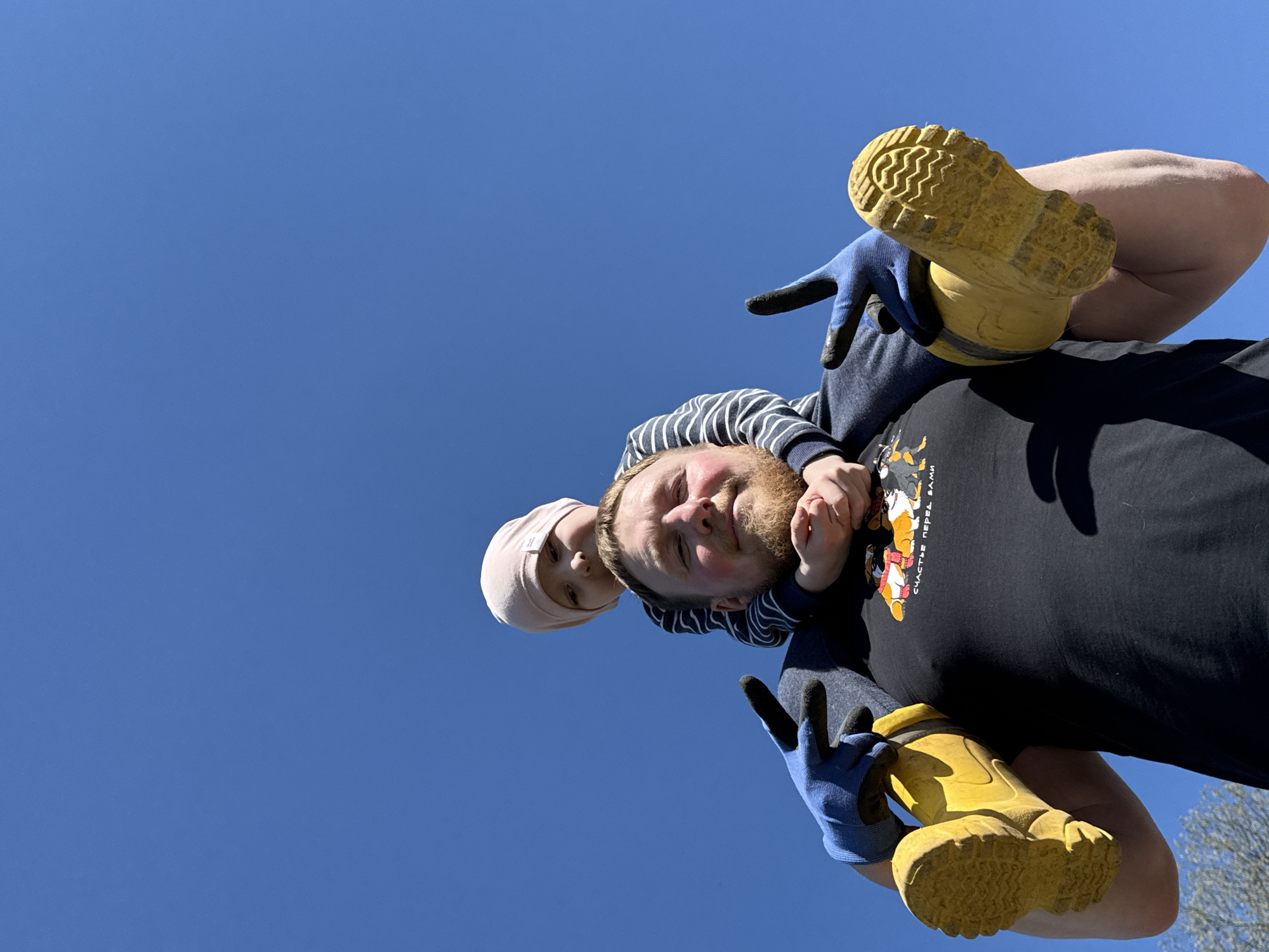 A low-angle shot depicts a man with a fair complexion and a reddish beard, carrying a young child on his shoulders. The child, wearing a pink beanie, is positioned behind the man's head. The man is wearing a black t-shirt, and blue work gloves, and is holding a pair of yellow rubber boots. His fingers are extended in a "peace sign" gesture. The background is a clear blue sky. The print on the shirt includes Russian text "СЧАСТЬЕ ПЕРЕД ВАМИ" which roughly translates to "Happiness is before you". Based on the text, the scene may potentially be located in Russia. The city of Nizhnyaya Chermoda, Russia is not verifiable based on available image details.
