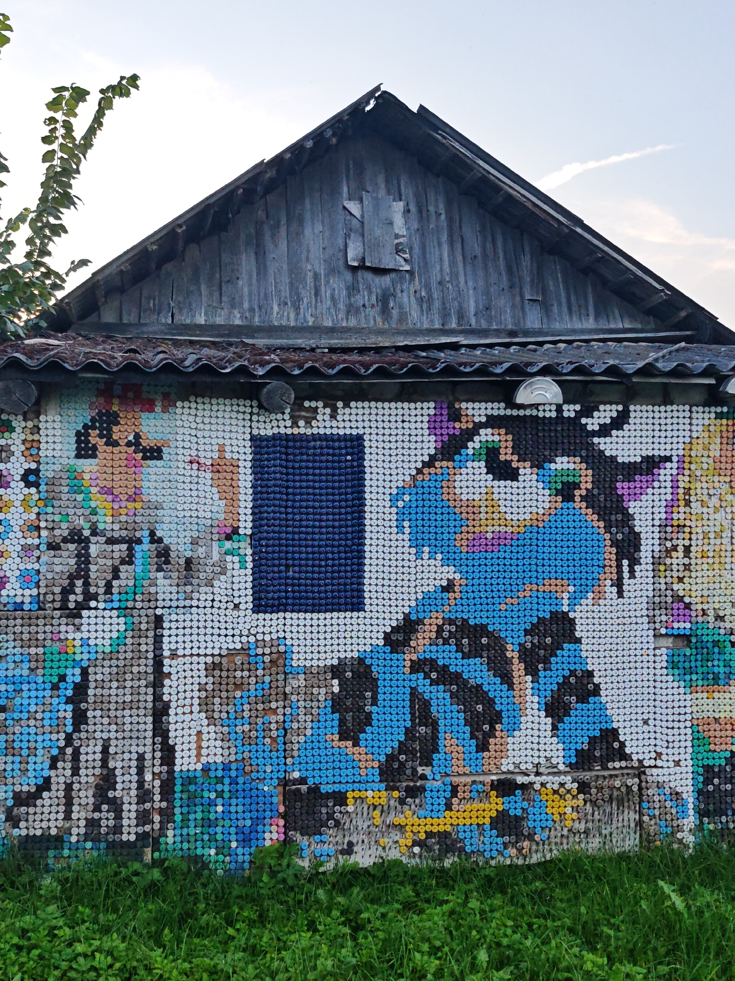 The image shows the exterior of a wooden structure, likely a shed or an outbuilding, in Валожынскі сельскі Савет, Belarus. The walls of the structure are extensively decorated with a mosaic made from colorful plastic bottle caps. The artwork depicts cartoon-like figures, including what appears to be a cat with large green eyes wearing a blue and black striped outfit. A dark blue rectangular shape, possibly representing a window, is incorporated into the mosaic on the right side of the structure. The roof is made of corrugated material and is in a state of disrepair, with weathered wooden planks forming the gable end. A tree with green leaves is visible on the left side, reaching towards the sky. The ground in front of the structure is covered with lush green grass. The sky is partly cloudy.