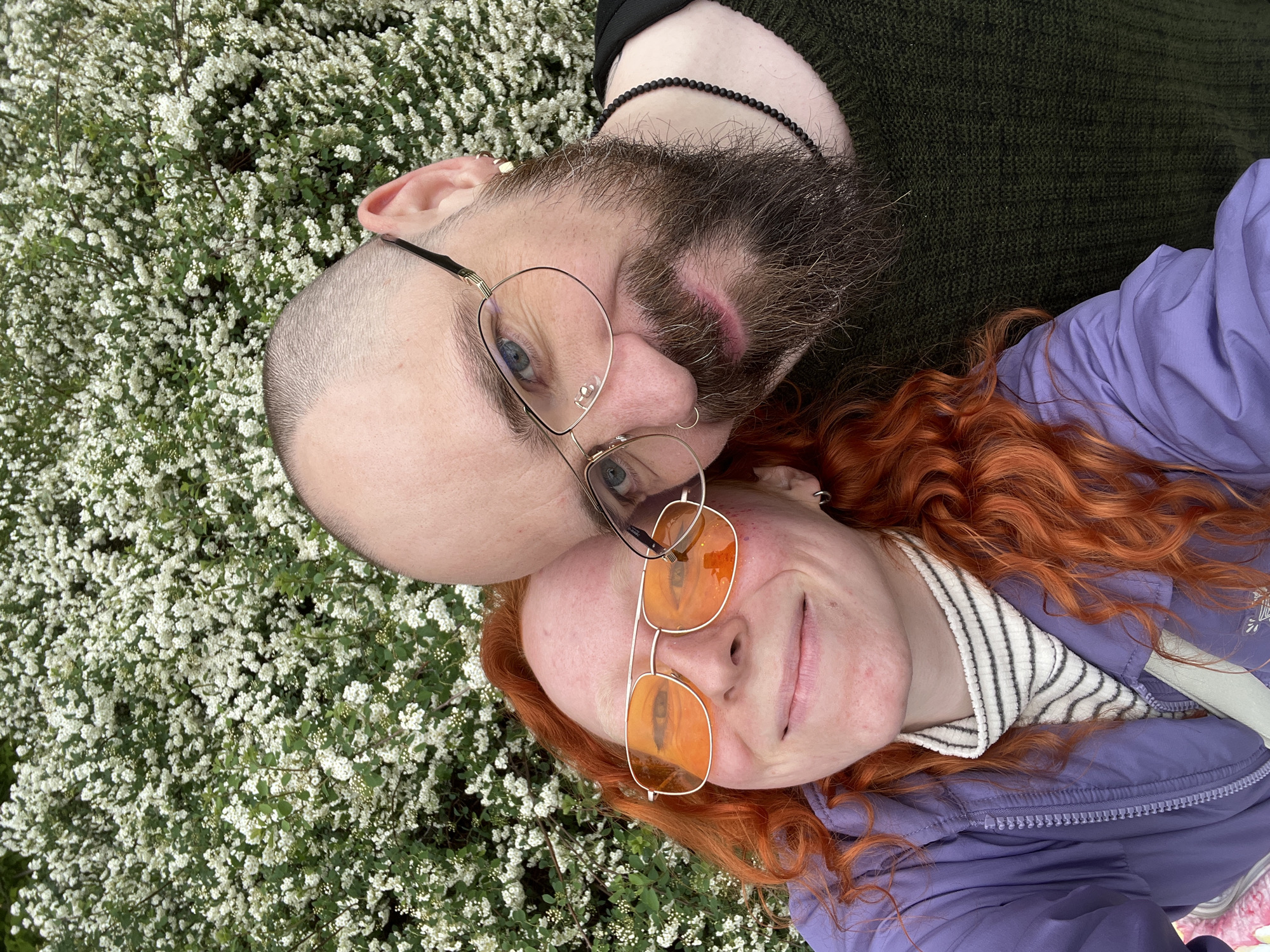 This close-up selfie captures two individuals lying down amidst abundant white flowering bushes, likely in a park or garden in Belgrade, Serbia. The person on the left has a shaved head, a prominent beard, and wears clear-rimmed glasses, revealing blue eyes. They are dressed in a dark green knitted sweater and wear a black beaded necklace. Resting beside them is a person with long, wavy, bright red hair, sporting orange-tinted aviator-style glasses. This individual is wearing a purple jacket with a striped inner collar and has a small silver hoop earring. Both are looking up towards the camera with relaxed, content expressions; the person with red hair has a gentle smile. The background is dominated by a dense array of small white flowers and green foliage, suggesting a vibrant spring or early summer setting. The bright, natural lighting indicates it is daytime. No visible text is present in the image.