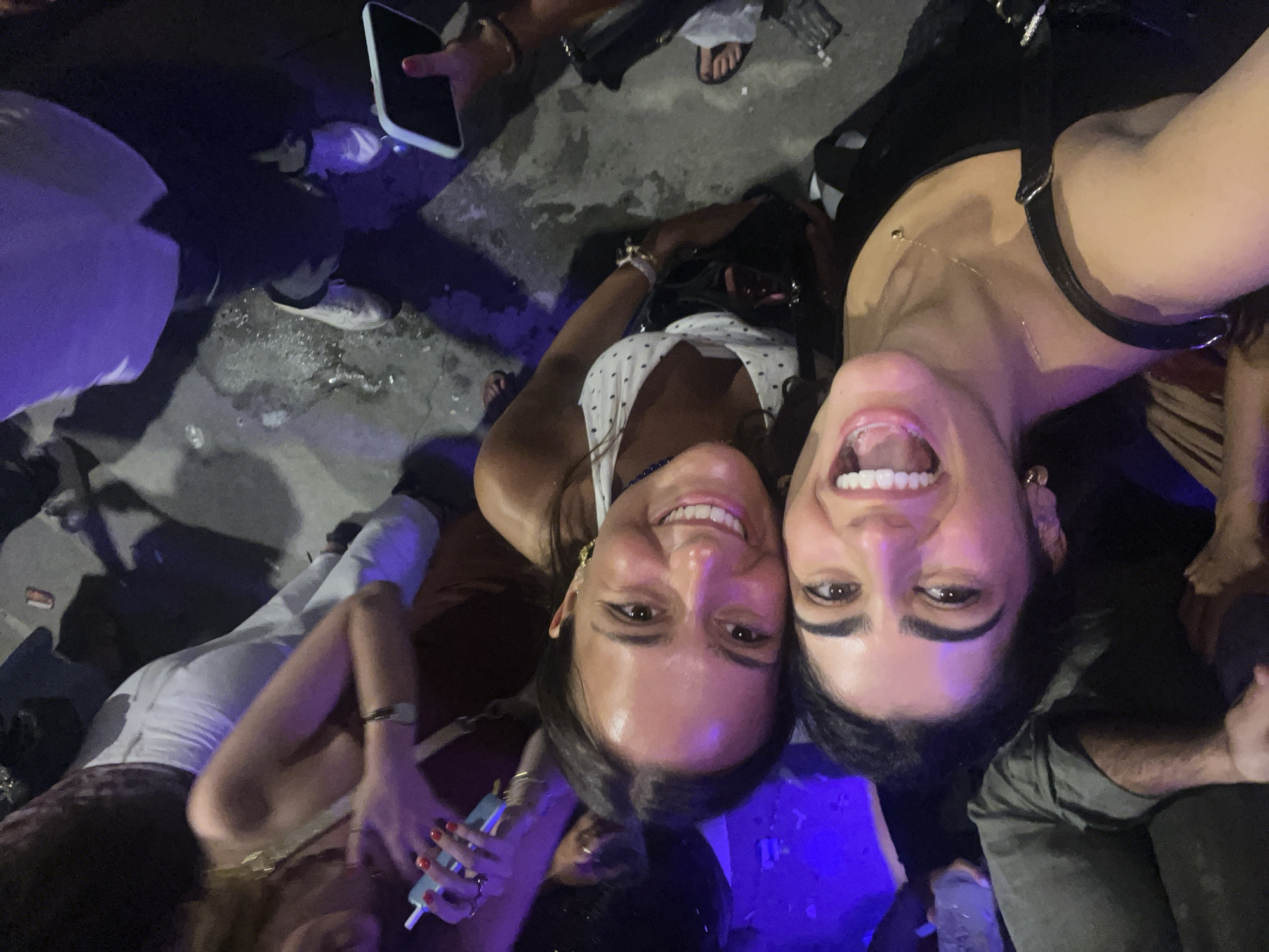 Two young women are taking a selfie outdoors at night, possibly in Quarteira, Portugal. The woman on the left has dark hair and is wearing a black top. She is smiling widely with her mouth open. The woman on the right has brown hair and is wearing a white top with a polka dot pattern. She is also smiling. They are surrounded by other people, some of whom are blurred in the background. The ground appears to be concrete or asphalt. There are some discarded items on the ground, and the lighting suggests an urban setting at night.