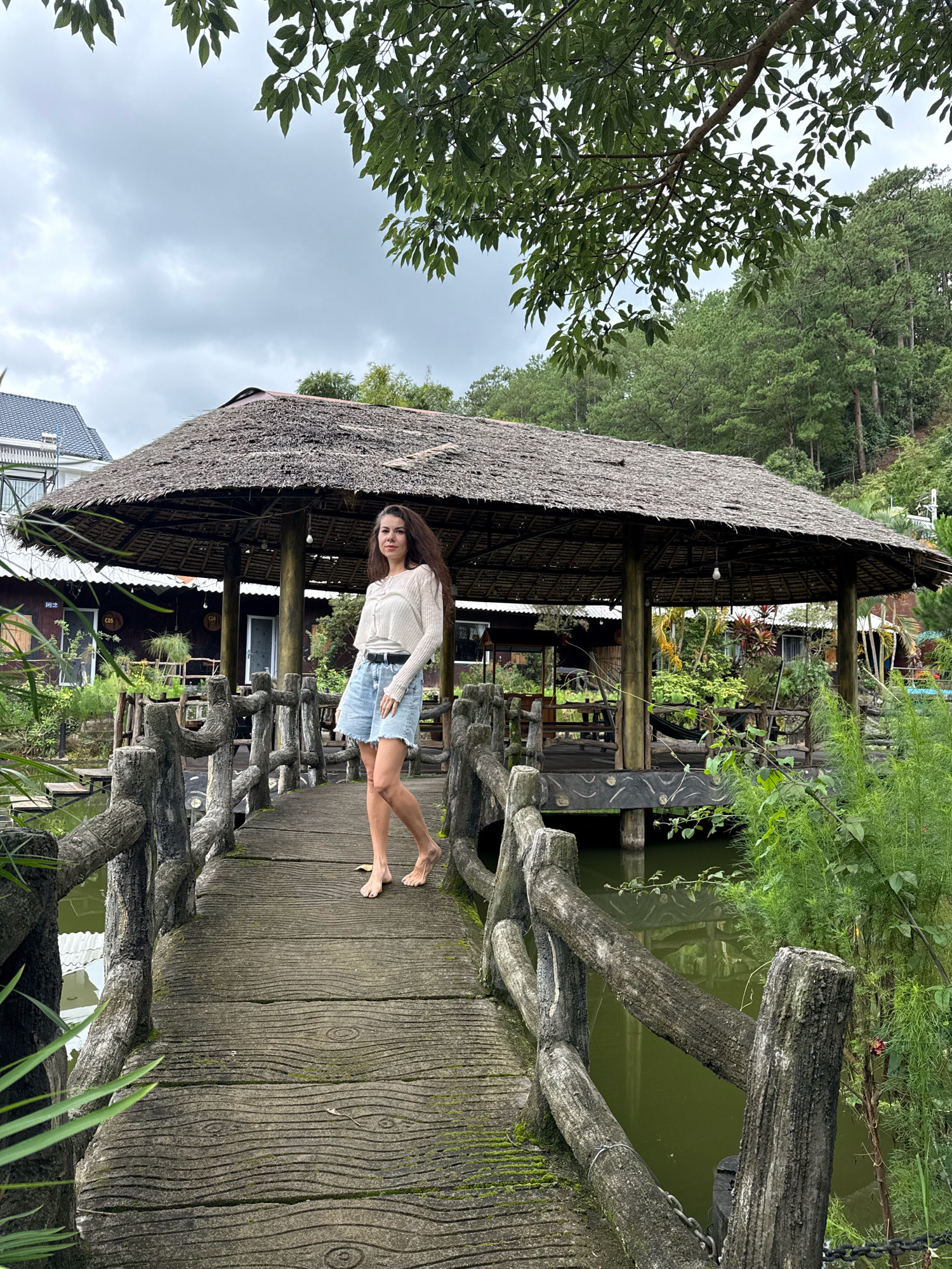 A full-shot, eye-level, outdoor, natural-light photograph captures a woman standing on a concrete bridge over a body of water, at a tropical resort in Hiệp Thạnh, Vietnam. The woman, who appears to be in her late 20s, has long, dark hair and is wearing a cream-colored, long-sleeved, unbuttoned shirt over a black t-shirt, denim shorts, and a black belt. She is barefoot. She stands in the middle of the bridge, facing forward, with her right leg bent and her left leg extended back.
The bridge is made of rough-textured concrete with a wood-grain pattern. It has a rustic wooden railing made of branches and concrete posts. The bridge leads towards a thatched-roof pavilion with wooden posts and open sides, where seating is visible. Lush green vegetation surrounds the pavilion and the bridge, with trees and plants growing on the bank of the water. In the background, there are more trees on a hillside and the roof of a building. The sky is overcast and gray.